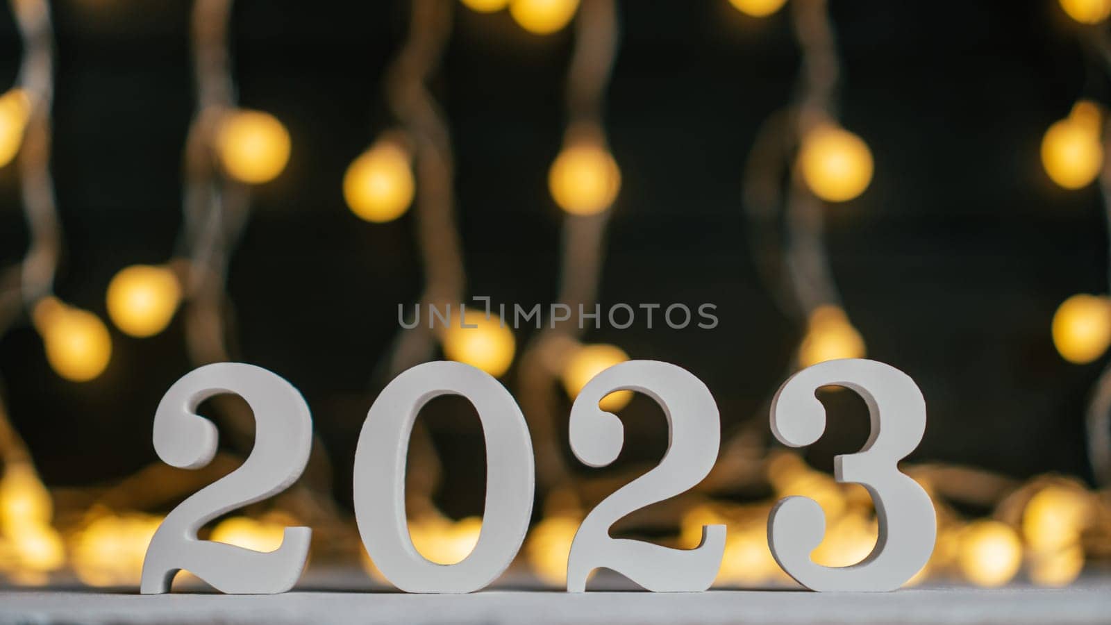 Happy New Year 2023. White wooden numbers 2023 stand on white surface against background of garland light bulbs. Merry Christmas. Copy space