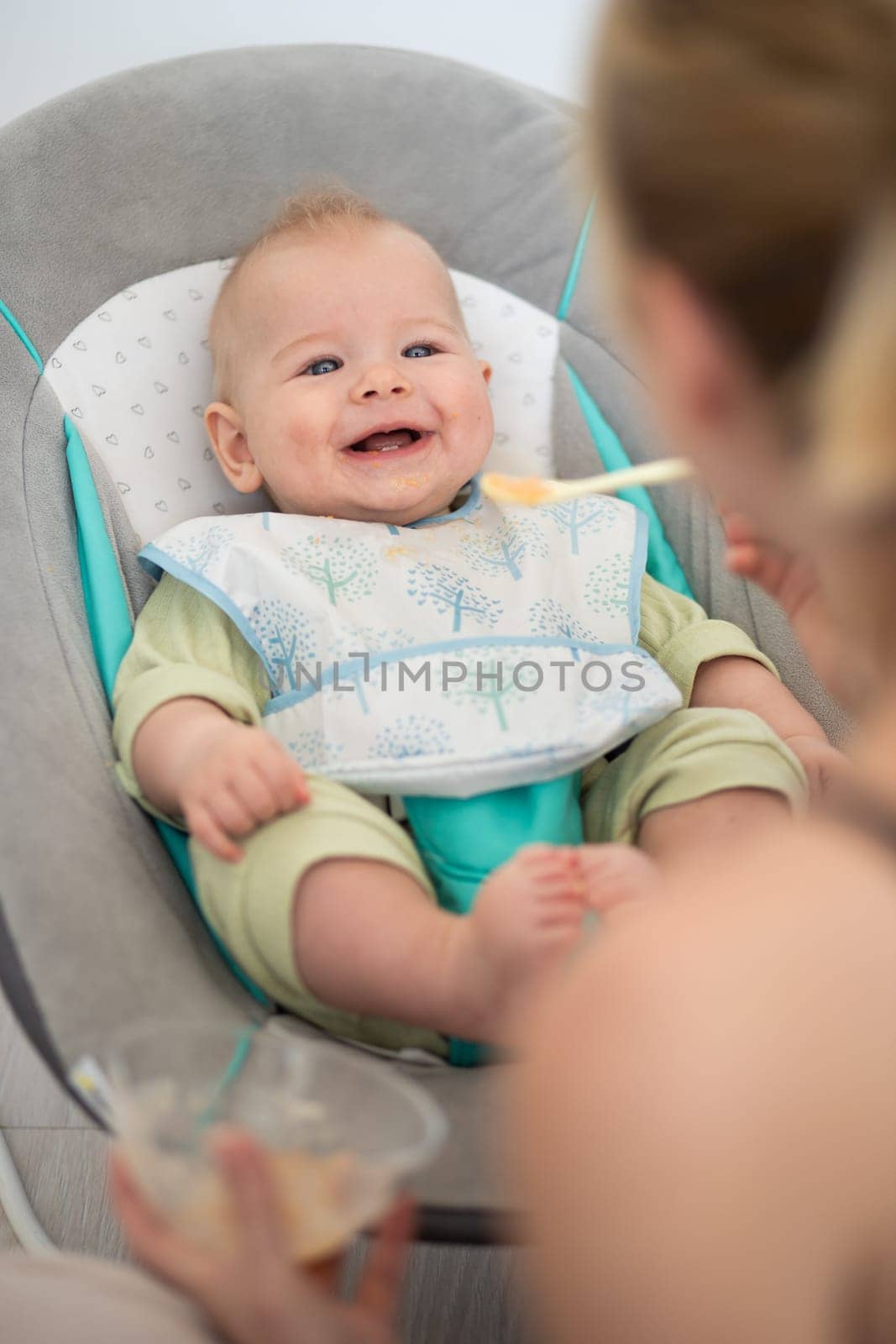 Mother spoon feeding her baby boy child in baby chair with fruit puree. Baby solid food introduction concept