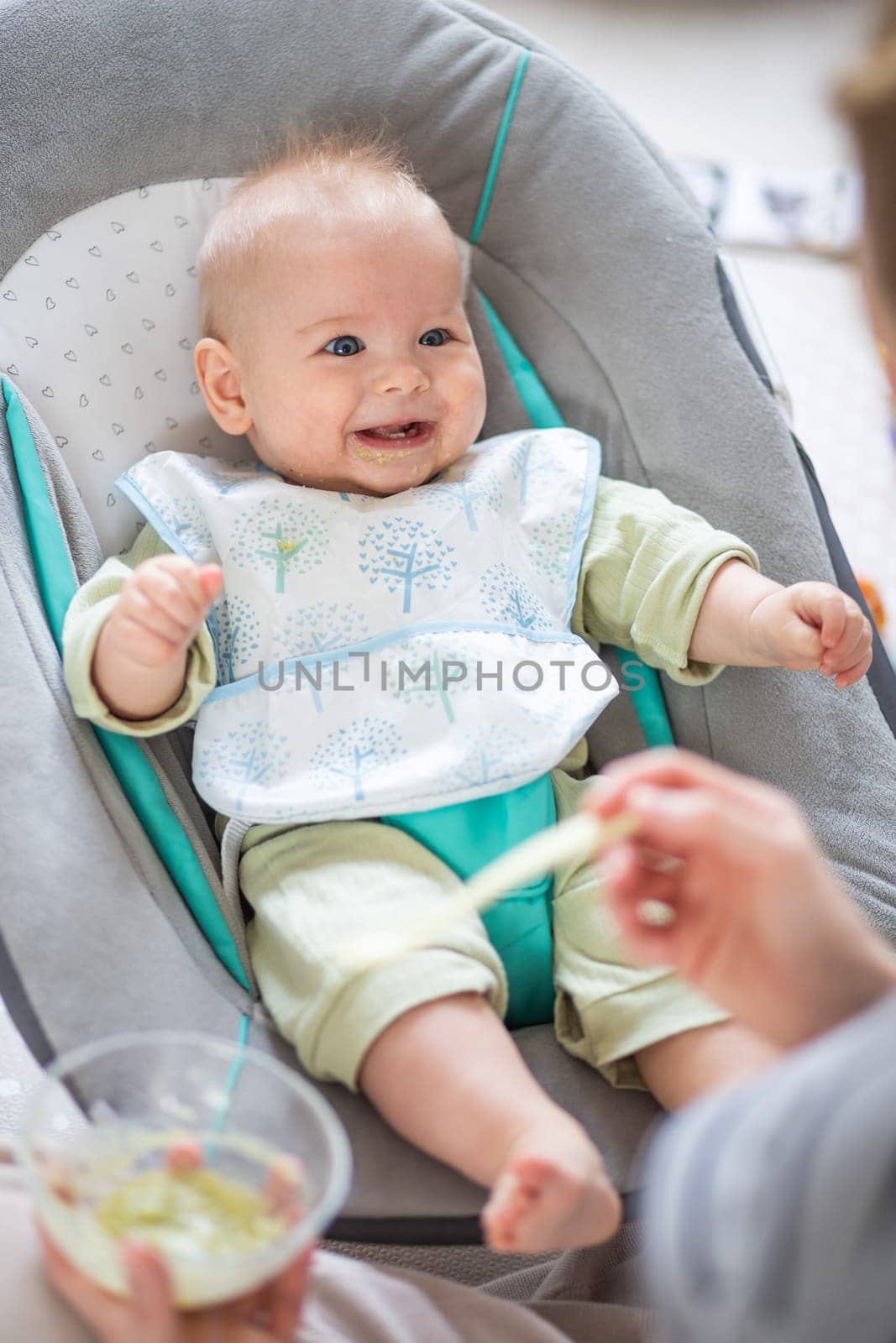 Mother spoon feeding her baby boy child in baby chair with fruit puree. Baby solid food introduction concept