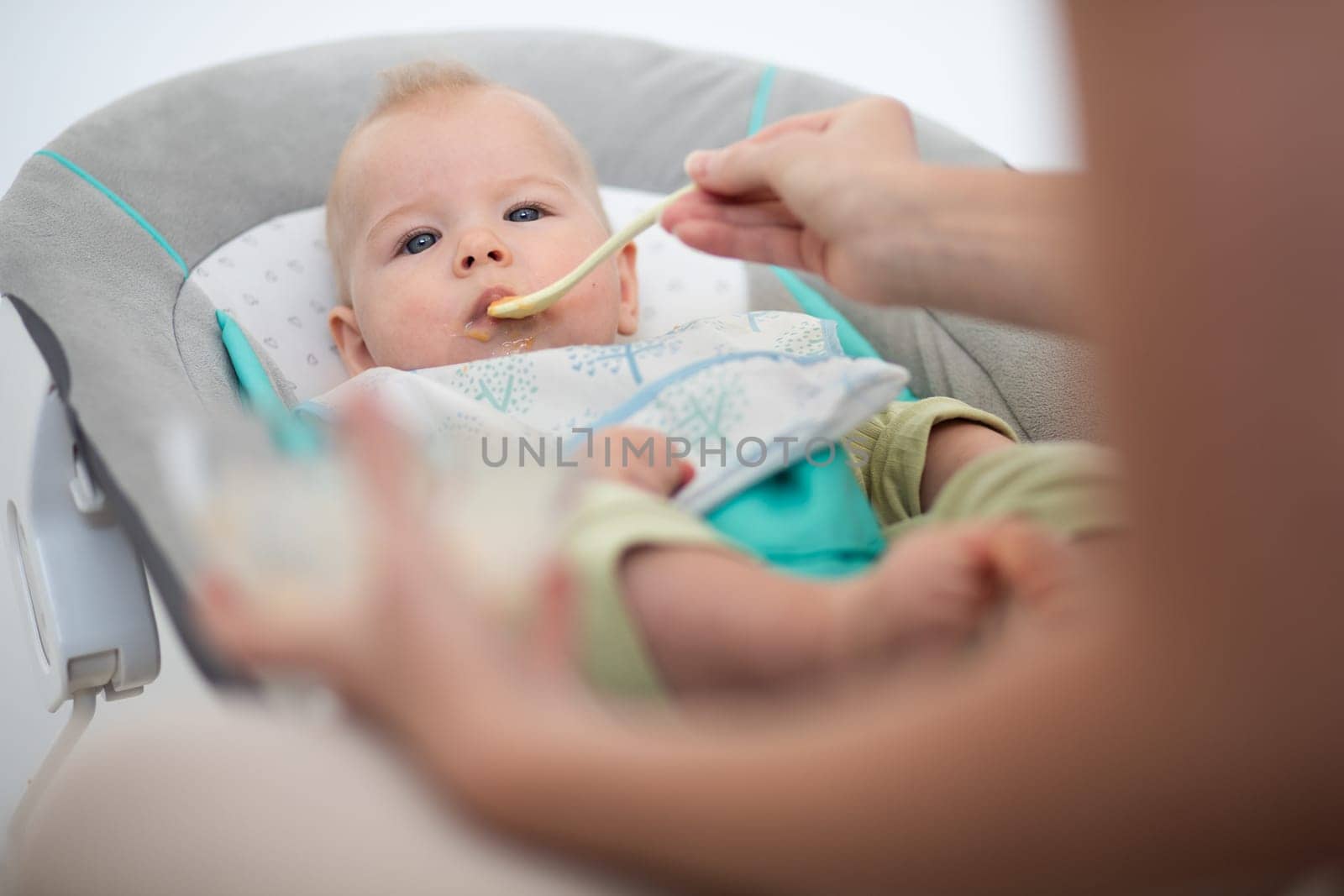 Mother spoon feeding her baby boy child in baby chair with fruit puree. Baby solid food introduction concept