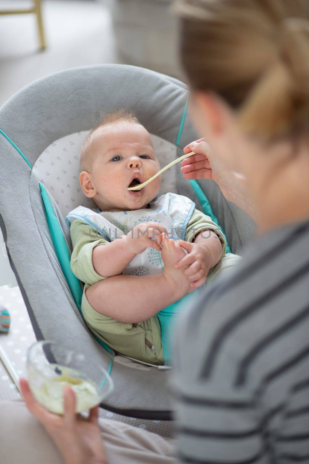 Mother spoon feeding her baby boy child in baby chair with fruit puree. Baby solid food introduction concept
