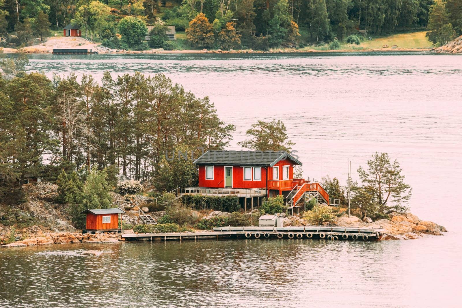 Sweden. Many Beautiful Red Swedish Wooden Log Cabin House On Rocky Island Coast In Summer. Lake Or River Landscape.