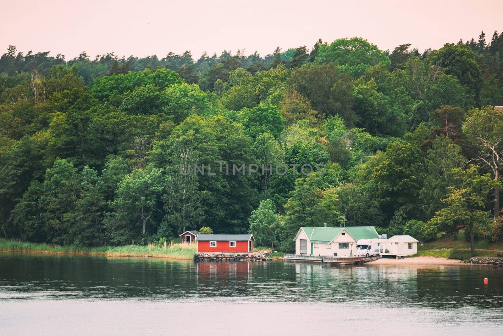 Sweden. Beautiful Red Swedish Wooden Log Cabin House On Rocky Island Coast In Summer. Lake Or River And Forest Landscape.