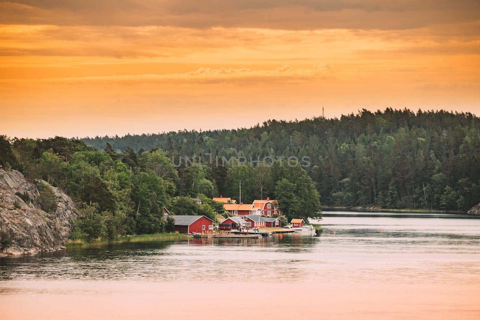 Sweden. Many Beautiful Red Swedish Wooden Log Cabins Houses On Rocky Island Coast In Summer Evening. Lake Or River Landscape.