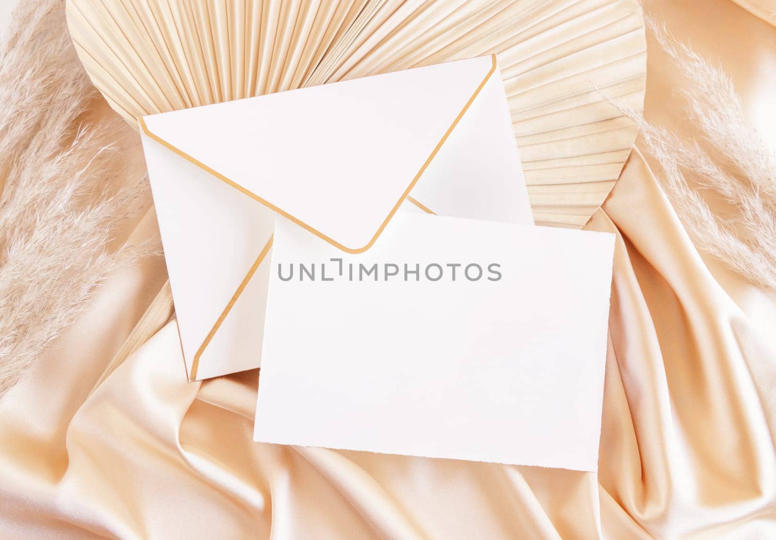 Card and envelope on beige satin fabric and dried palm leaf close up, mockup. Romantic Boho scene with horizontal paper card and envelope. Bohemian or close to nature greeting or wedding Invitation.