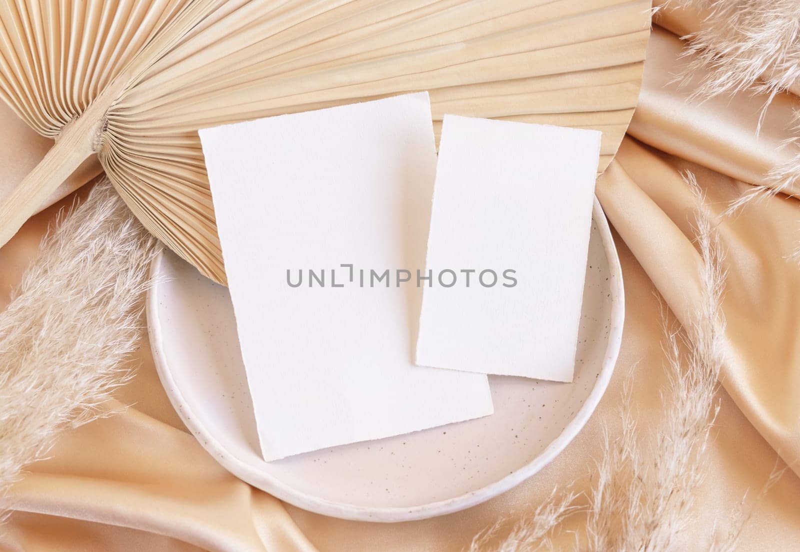 Blank cards on plate with dried palm leaf and beige silky fabric close up, mockup. Romantic scene with vertical paper cards. Bohemian pastel greeting or wedding Invitation .