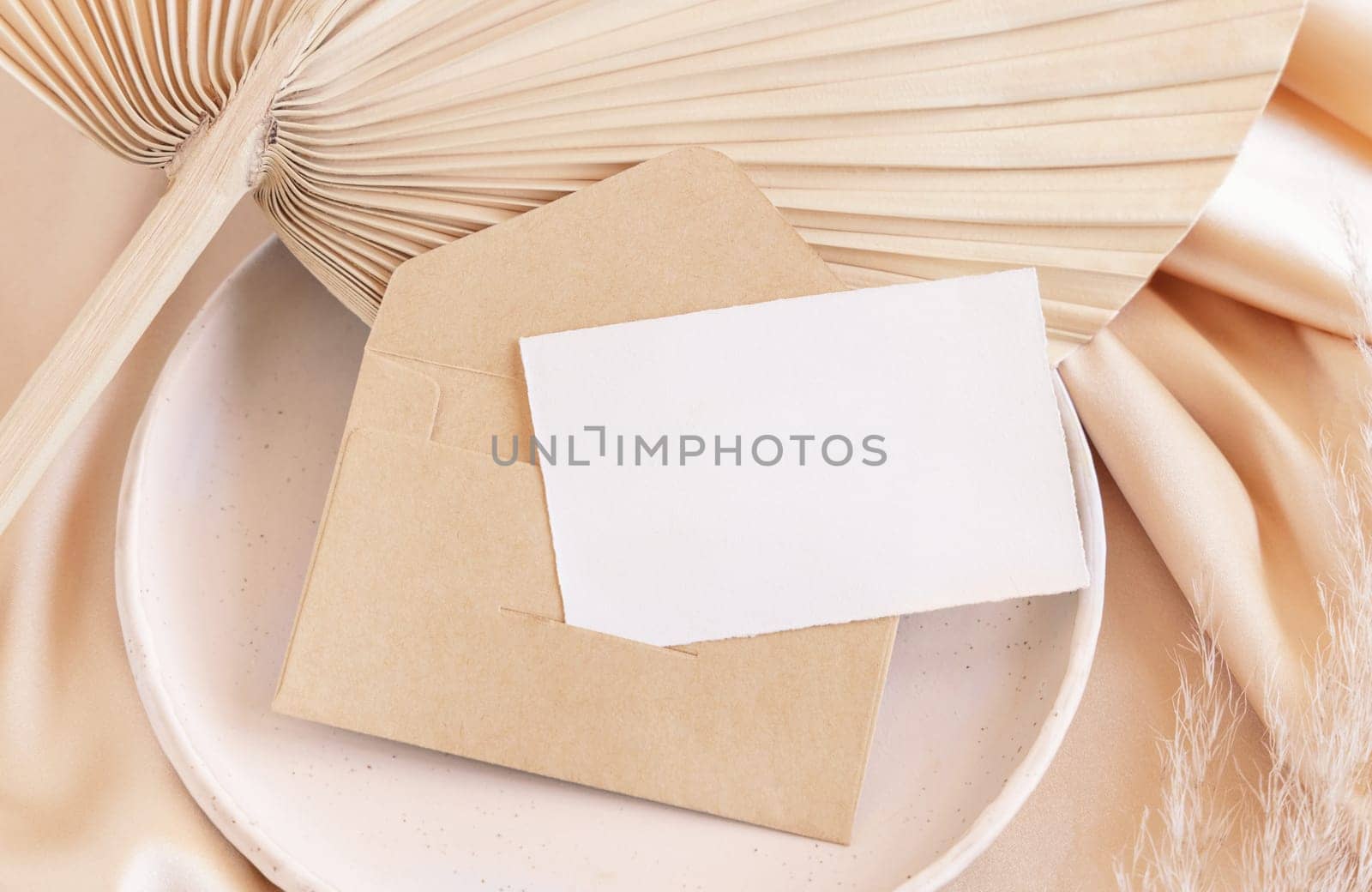 Blank card and envelope on plate with dried palm leaf and beige silky fabric close up, mockup. Romantic scene with horizontal paper card. Pastel greeting or wedding stationery.
