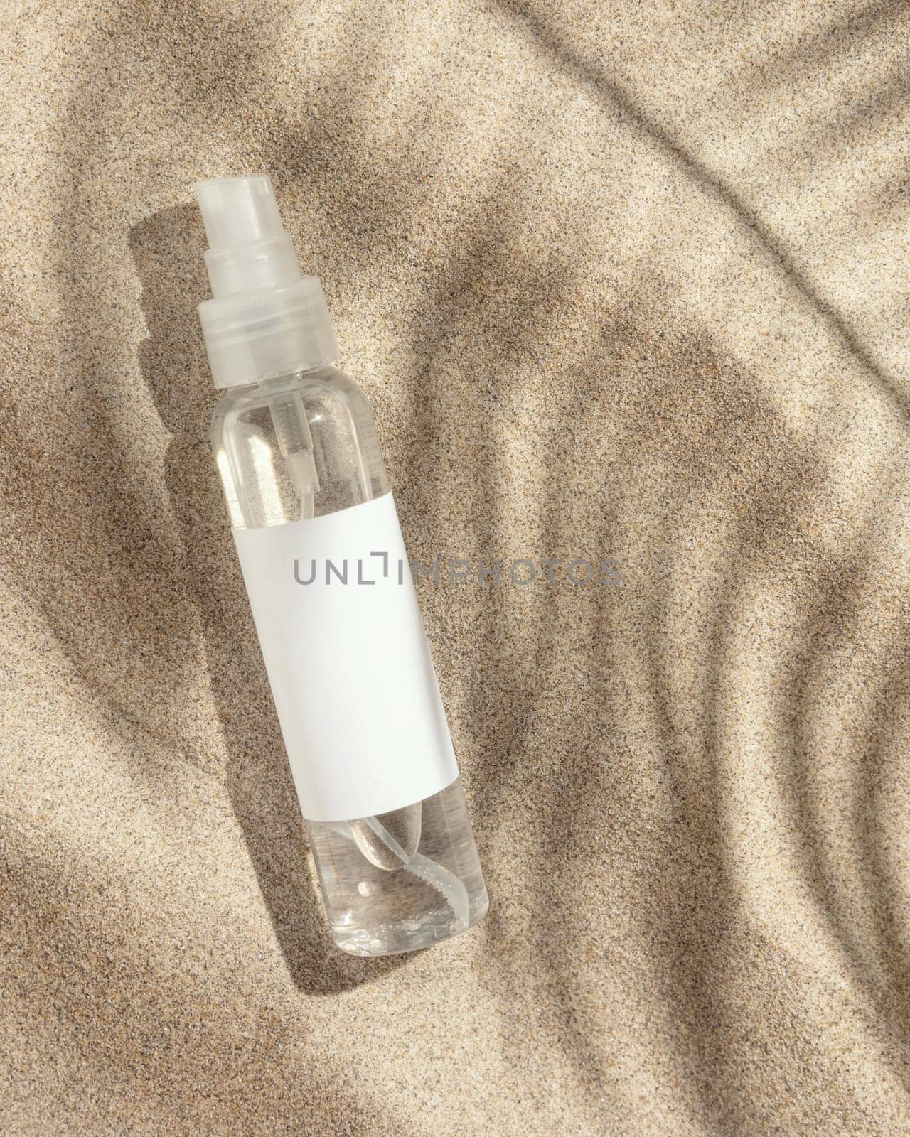 Cosmetic spray bottle with blank label on beige sand top view. Packaging mockup. Natural skincare product for beauty routine. Summer sunny day with palm leaf hard shadow, tropical vacation mood