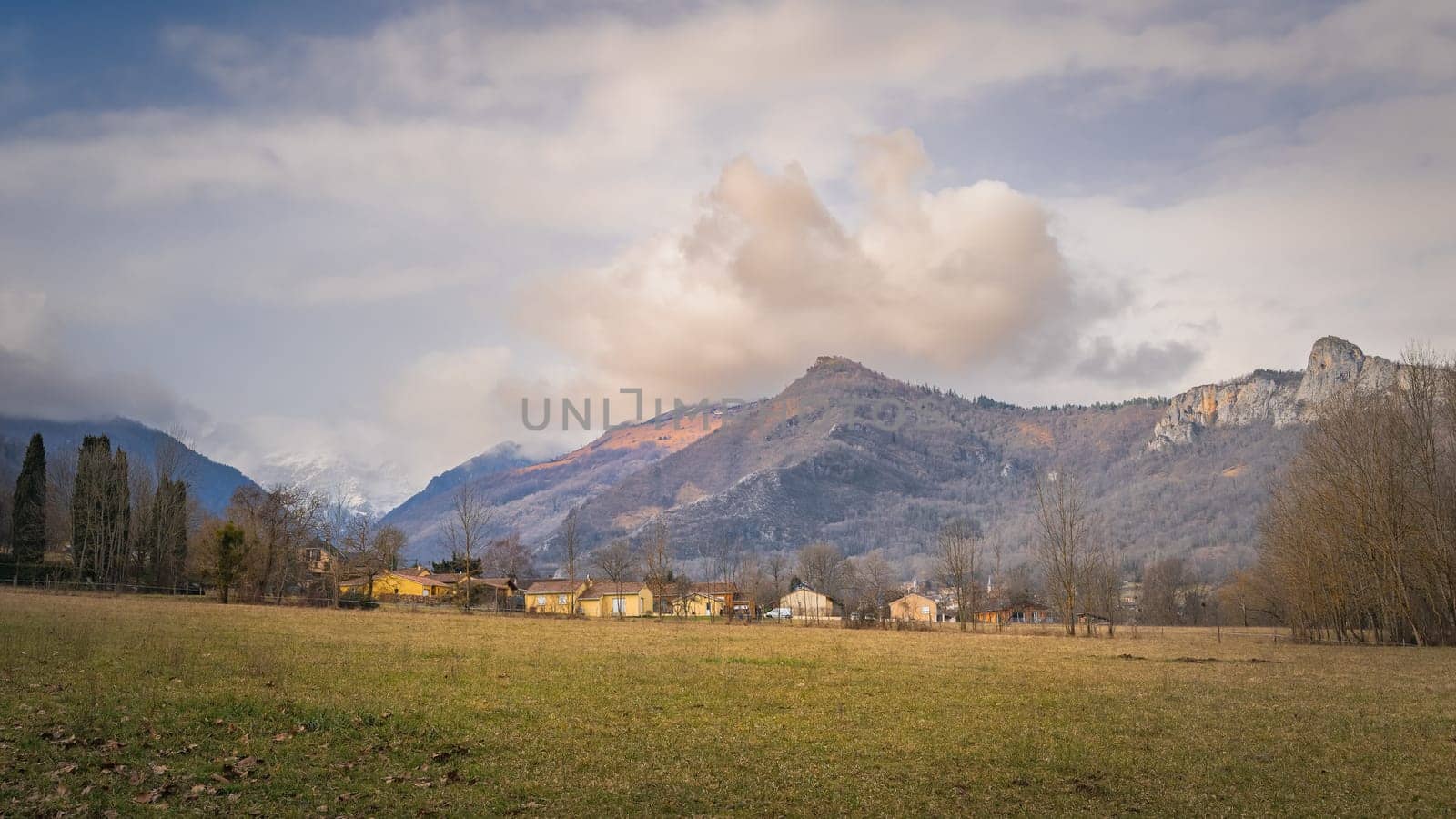 Surba town hiding behind trees at the foothill of Pyrenees Mountains with snowy peaks in background, Tarascon sur Ariege, France