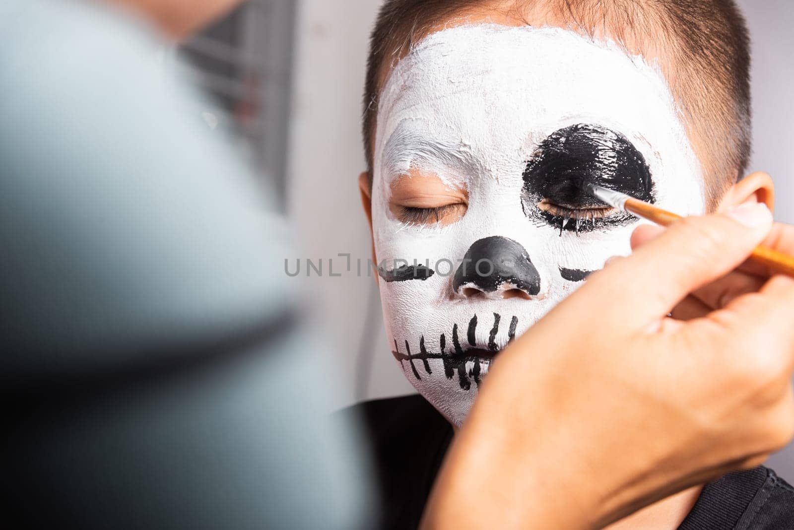 Asian mother and kid son getting makeup halloween face painting look like ghost, Portrait of woman applying color brush to child face for party, Happy halloween day concept