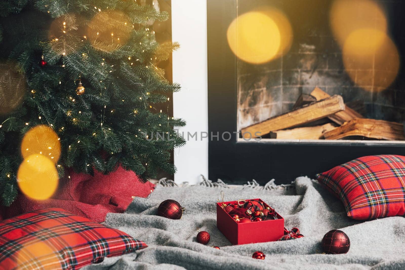 Box of Christmas balls laying on gray carpet or plaid ready for use for decorating and hanging on green Christmas fir tree with lights, garland in the living room near fireplace with wood inside