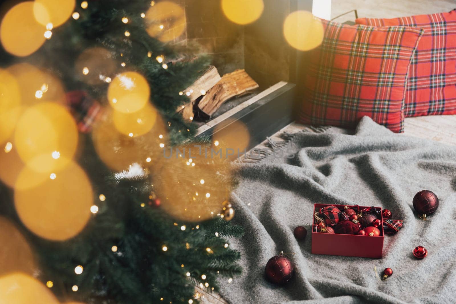 Box of Christmas balls laying on gray carpet or plaid ready for use for decorating and hanging on green Christmas fir tree with lights, garland in the living room near fireplace with wood inside