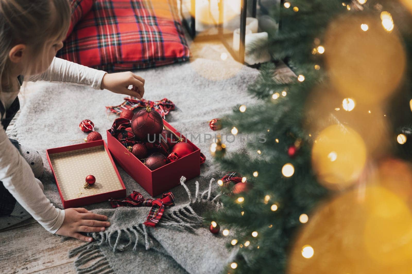 Little child girl decorates Christmas fir tree with red ornament balls. Small kid holding gift in hand before hanging it to tree with light, garland. Box with balls laying on floor with decorations