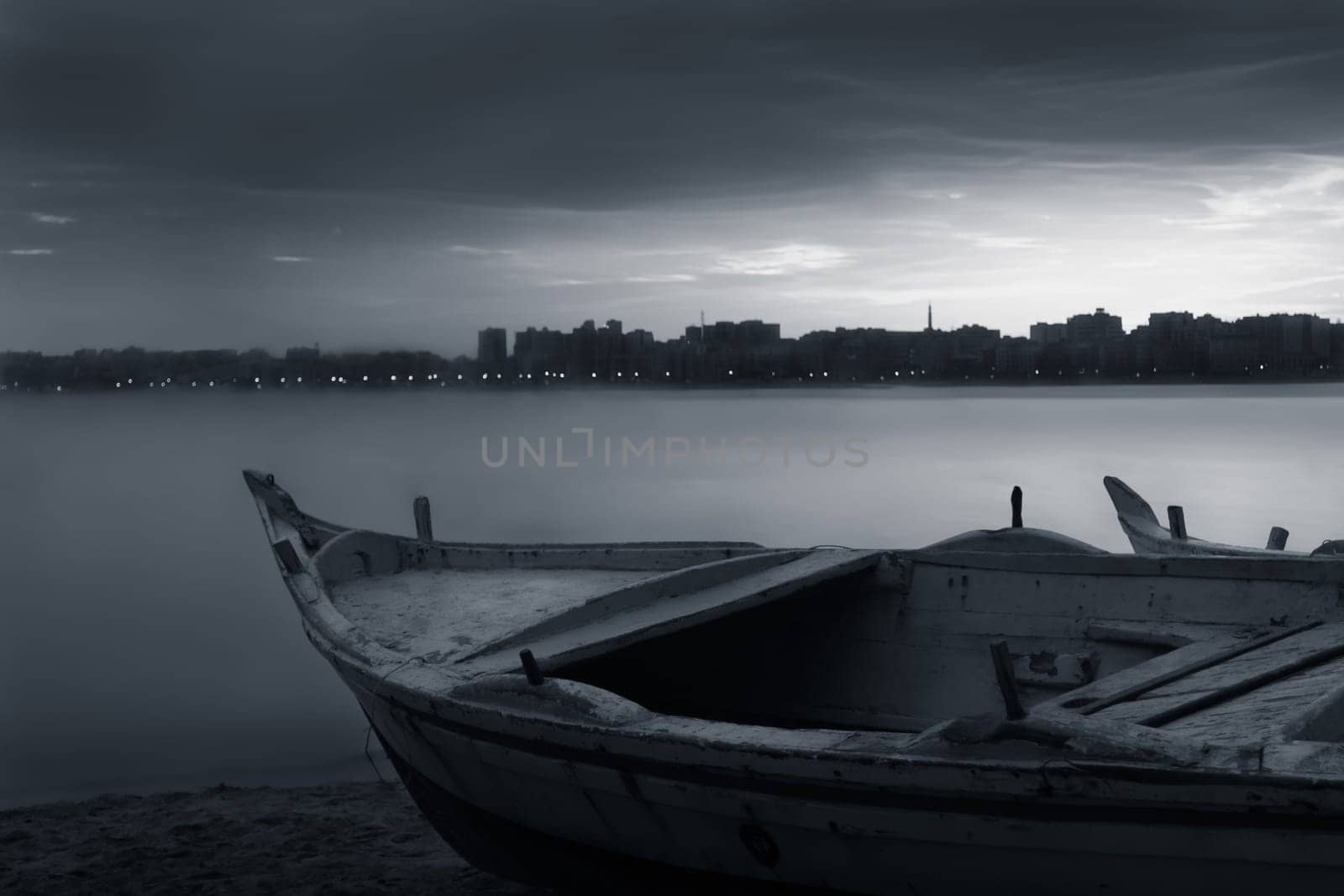 Black and white shot of fishing boat on the beach at sunrise with Egyptian Alexandria city skyline in far distance and cloudysky at sunrise