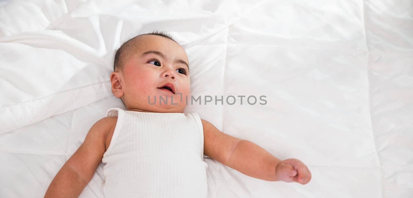 Asian smiling infant child at home, Cute little baby on bed under soft blanket, baby wearing bodysuit lying on white beedsheets in bedroom