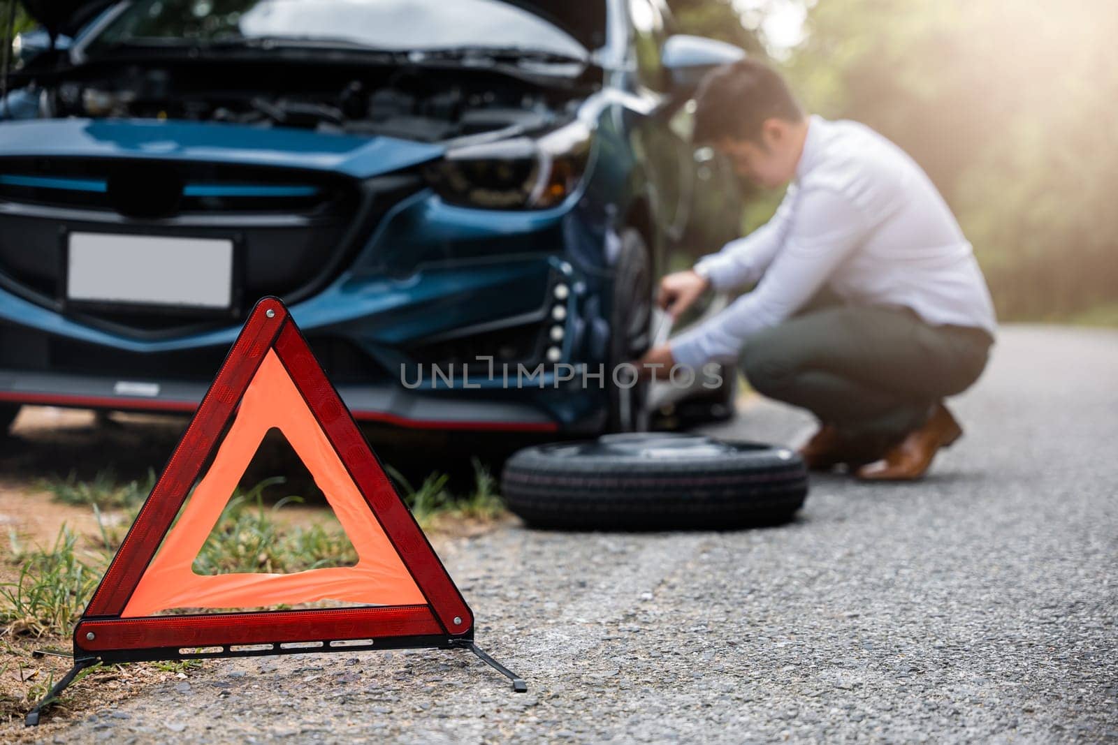 Asian businessman car broken has problems with wheel of his car during go to work in morning he changing replace rubber tire on wheels, business man have warning sign on road, problem transportation