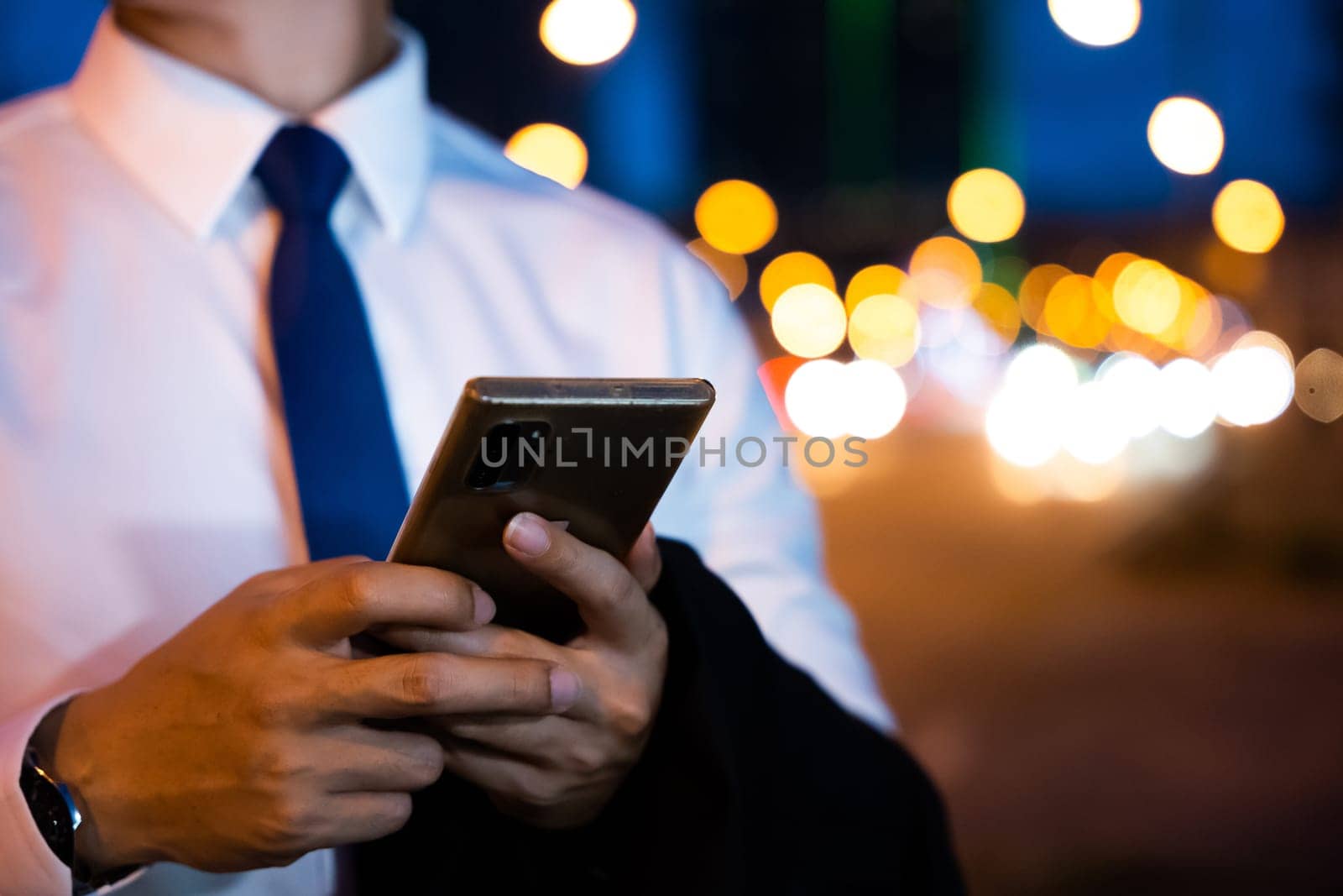 Portrait Asian businessman typing an sms message via smartphone after work near office at night city street, bokeh lights, young man walking and chatting with friends at social networks outdoor