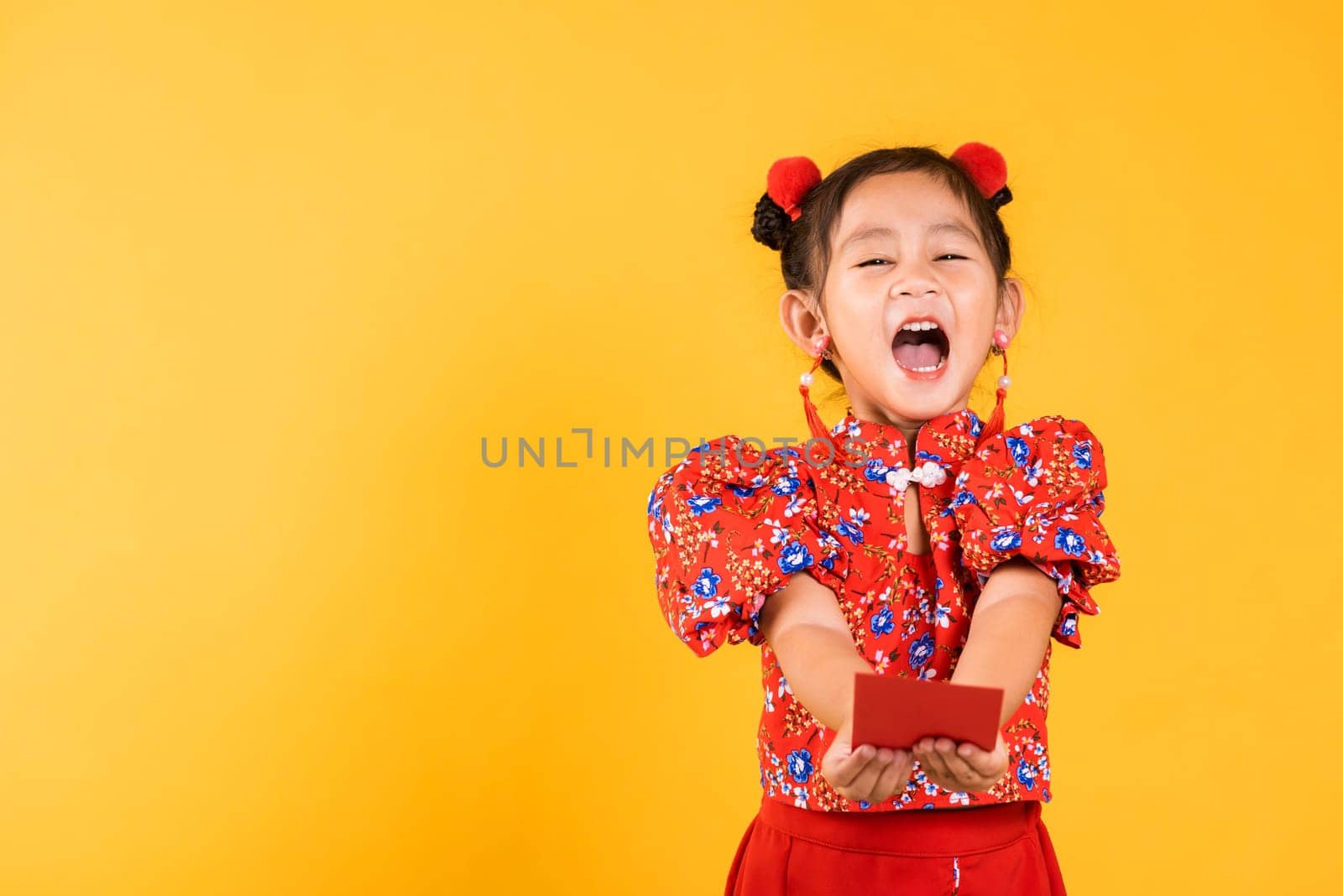 Chinese New Year. Happy Asian Chinese little girl smile wear red cheongsam handed angpao or red packet monetary gift, Portrait children in traditional dress, studio short isolated on yellow background
