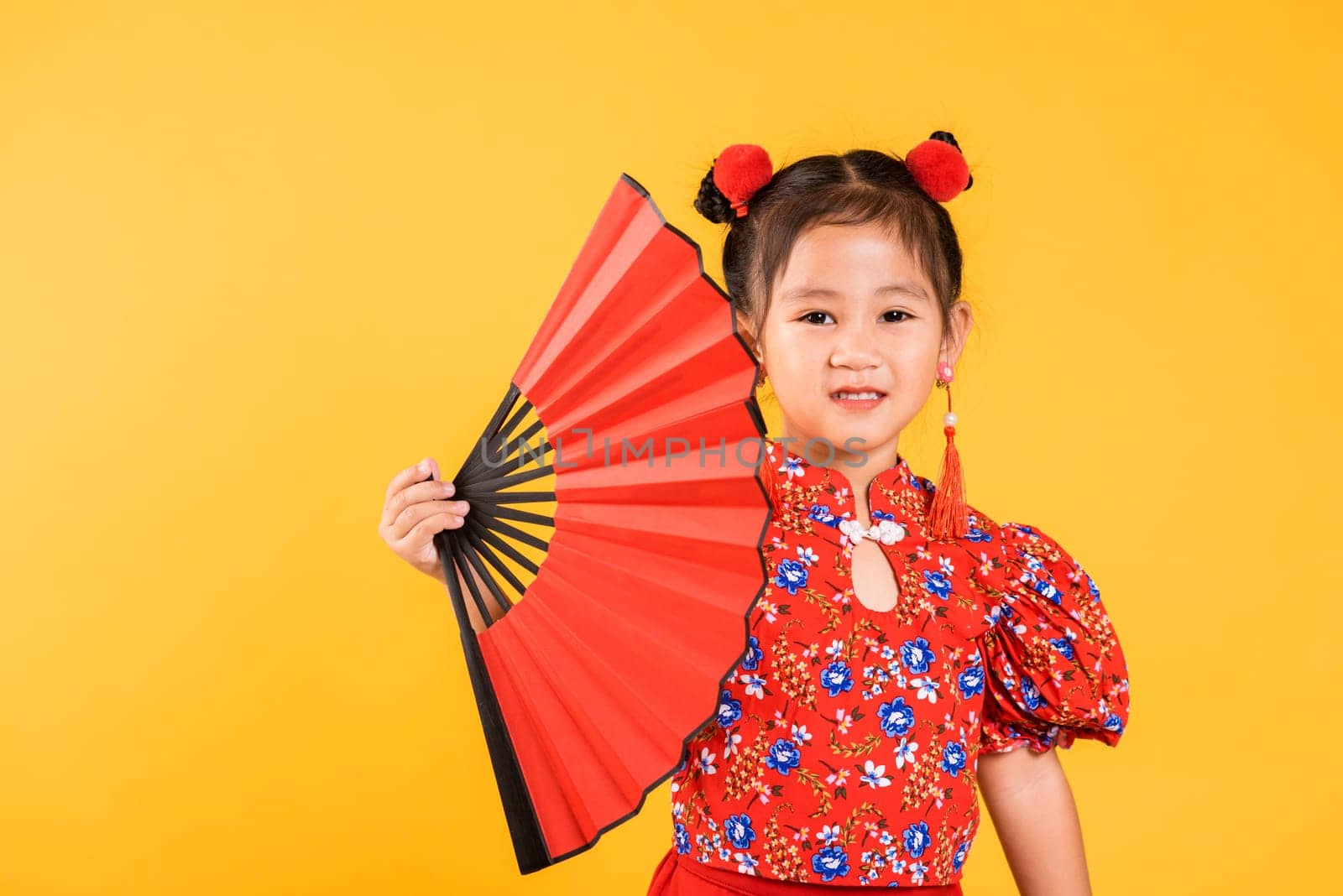 Chinese New Year. Happy Asian Chinese little girl smile wearing red cheongsam holding fan, Portrait children in traditional dress, studio short isolated on yellow background