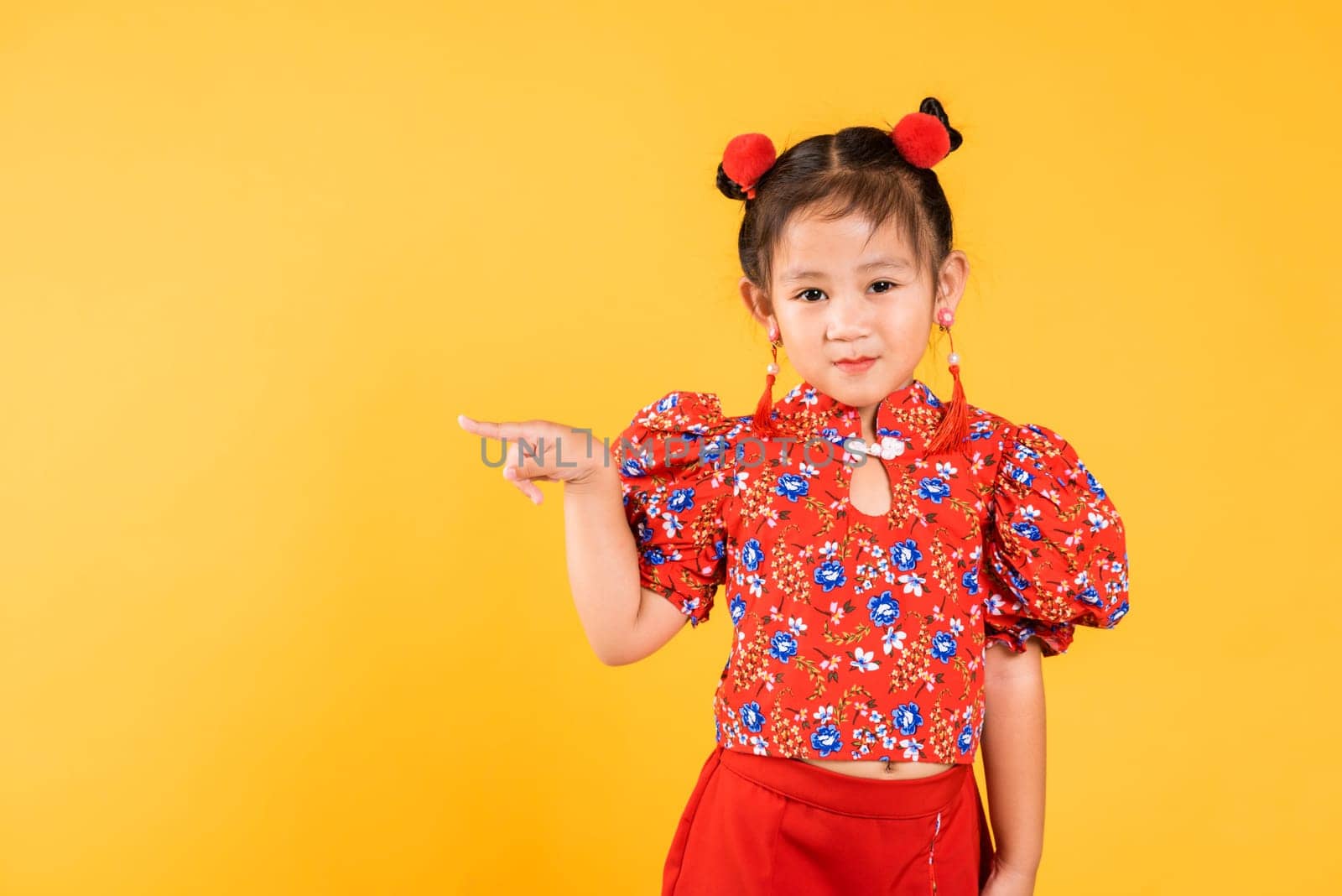 Happy Asian Chinese little girl wear red cheongsam qipao pointing finger to space, Portrait children in traditional dress point to empty, Chinese New Year, studio short isolated on yellow background