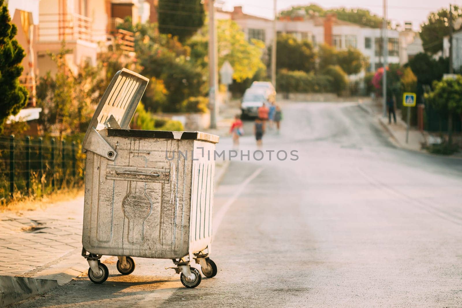 Metal Waste Container On Wheels In Street During Summer Sunny Evening. Container For Temporarily Storing Waste. Dustbin, Garbage Can, Trash Can.