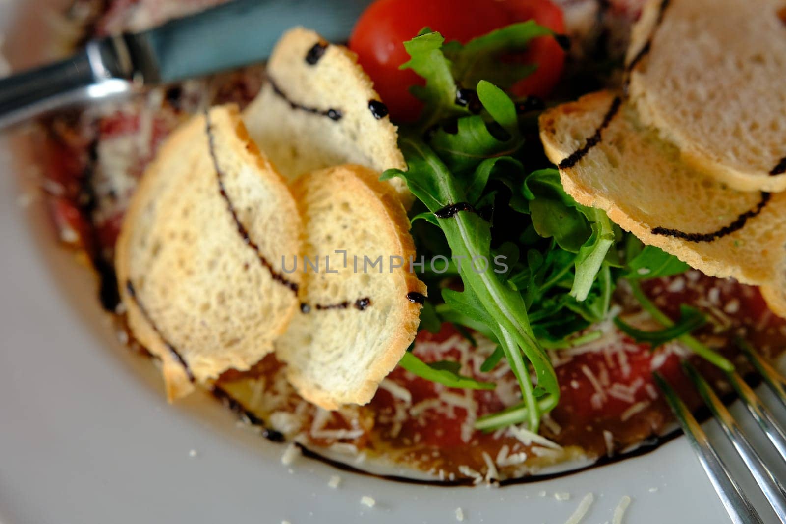 Beef Carpaccio cold appetizer with parmesan, tomatos and arugula on white plate with hardtack on dark background.