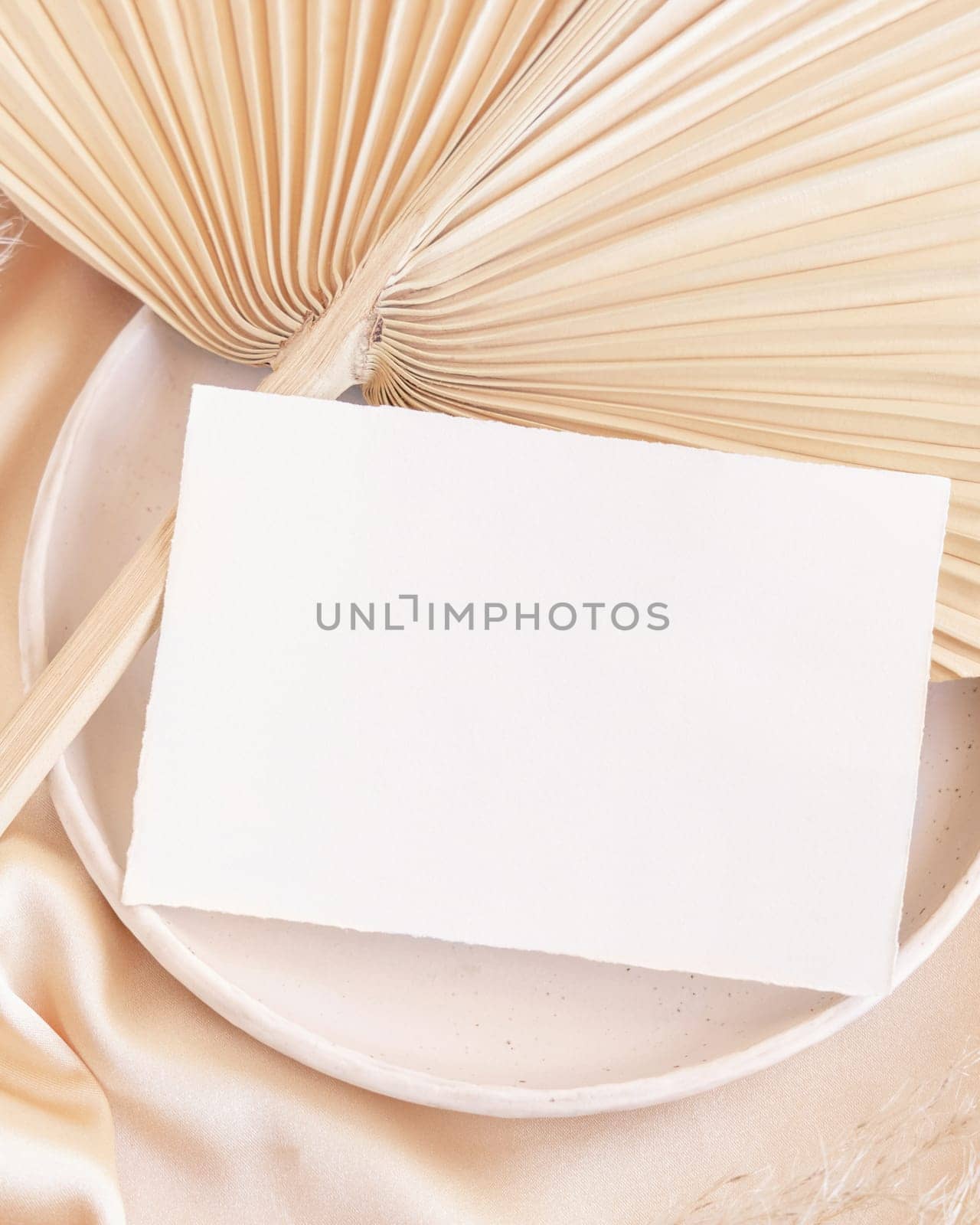Blank card on plate with dried palm leaf and beige silky fabric close up, mockup. Romantic scene with horizontal paper card. Bohemian pastel greeting or wedding Invitation .
