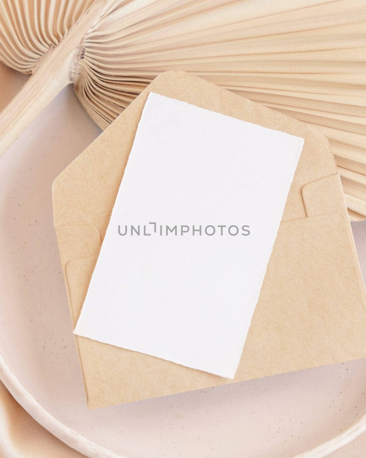 Blank card and envelope on plate with dried palm leaf and beige silky fabric close up, mockup. Romantic scene with vertical paper card. Pastel greeting or wedding stationery.