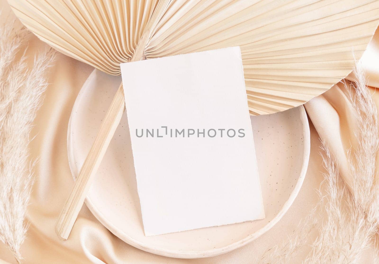 Blank card on plate with dried palm leaf and beige silky fabric close up, mockup. Romantic scene with vertical paper card. Bohemian pastel greeting or wedding Invitation .