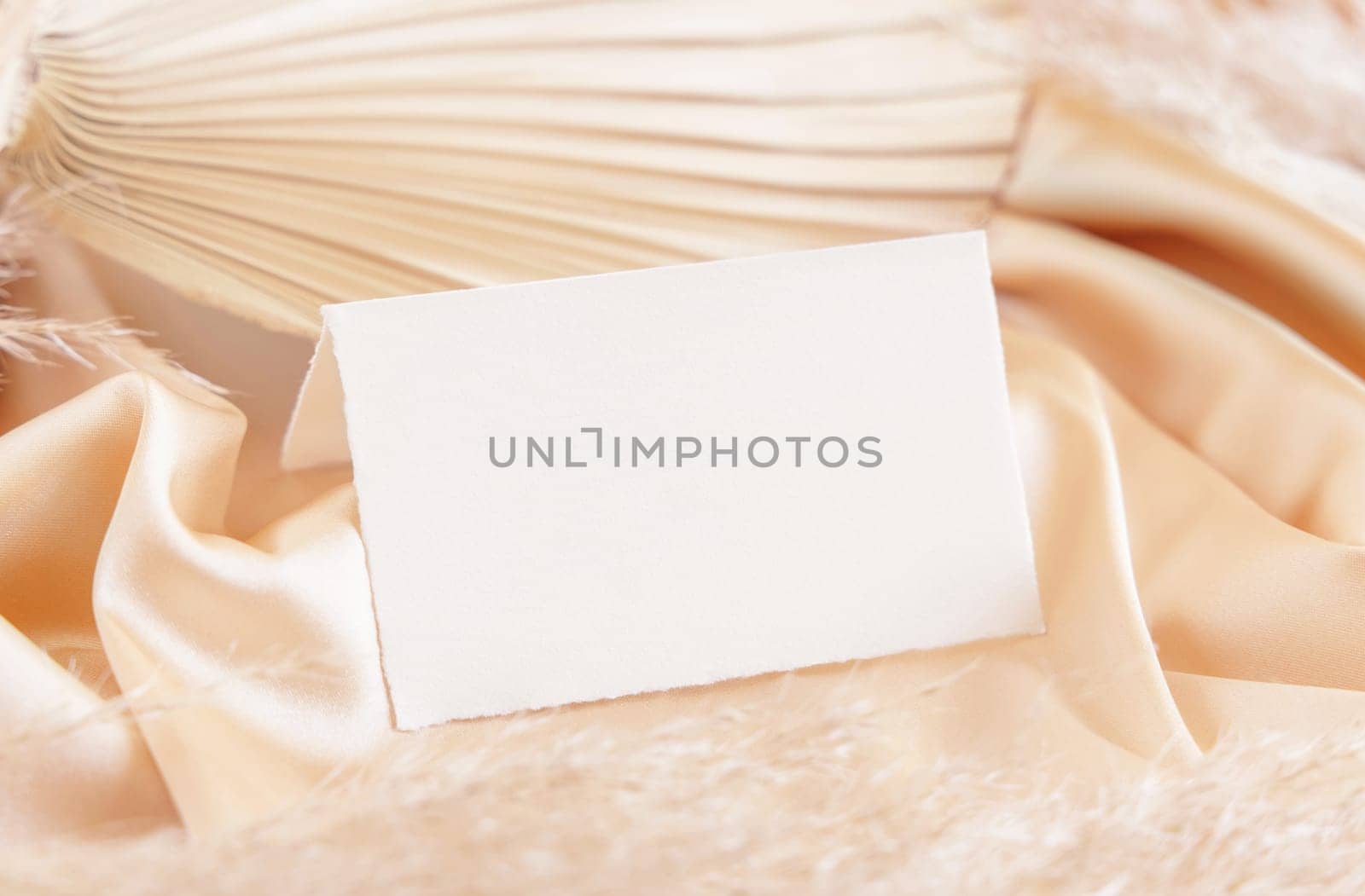 Folded place card on beige satin fabric and dried palm leaf close up, mockup. Boho scene with horizontal paper card. Bohemian or close to nature greeting or wedding Invitation. Romantic composition.