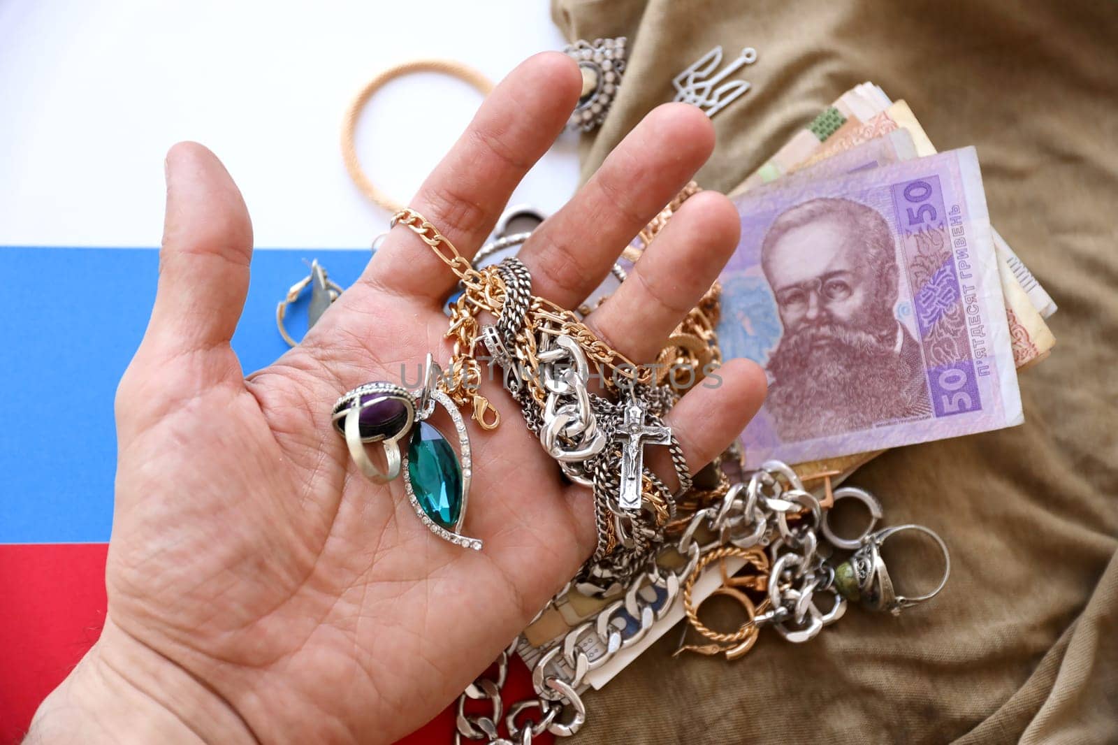 Marauders hand with bunch of stolen jewelry and money on military uniform cloth fabric and Russian flag. Looting by Russian soldiers in the Ukrainian cities during the Russian invasion