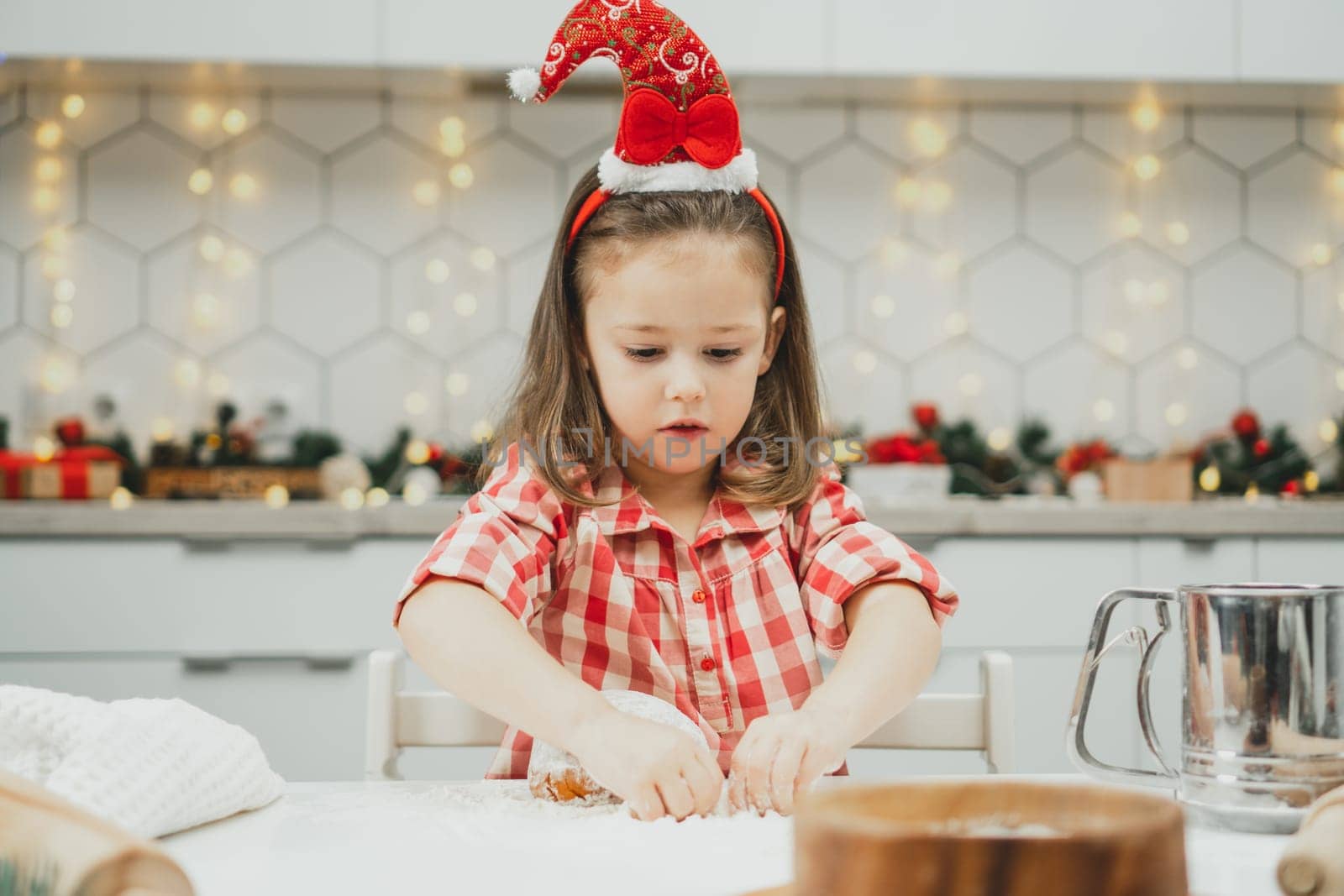 Little dark-haired girl 3 years old in red Christmas cap and checkered shirt prepares dough for gingerbread cookies in white Christmas decorated kitchen with garland lights. Merry Christmas, New Year