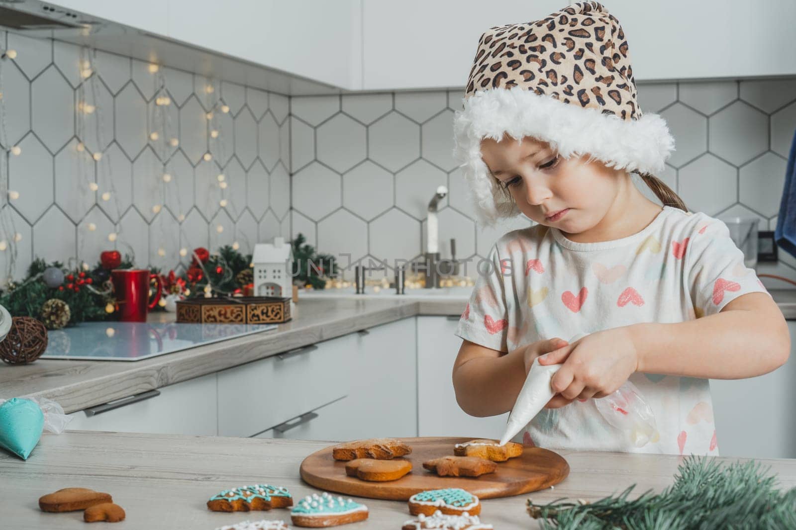 Little dark-haired girl 3 years old in Santa spotted cap decorate gingerbread cookies with icing in white Christmas decorated kitchen. Merry Christmas, Happy New Year