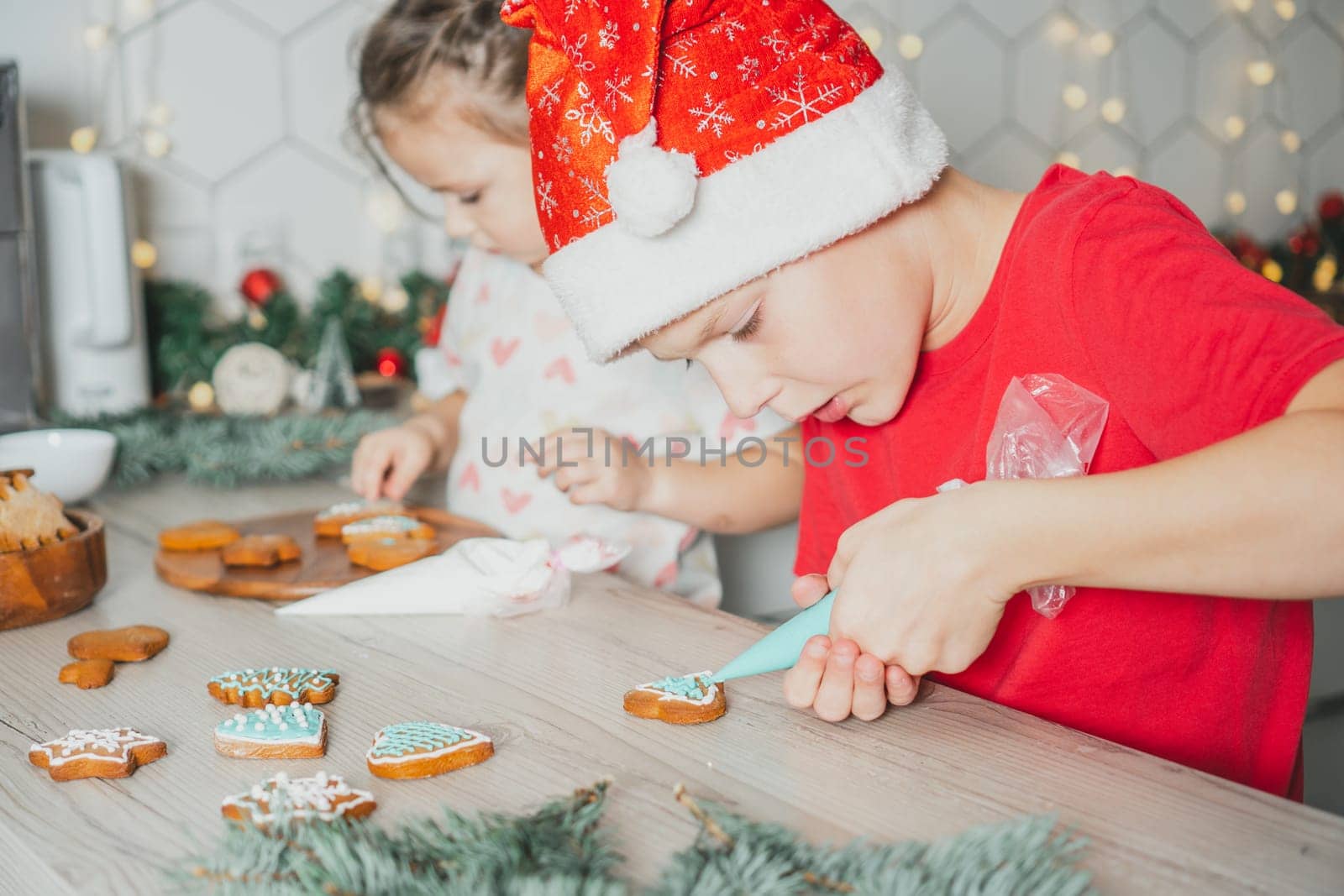 Little dark-haired girl 3 years old and boy 8 years old in red Santa hat decorate gingerbread cookies with icing. Siblings in white Christmas decorated kitchen. Merry Christmas, Happy New Year