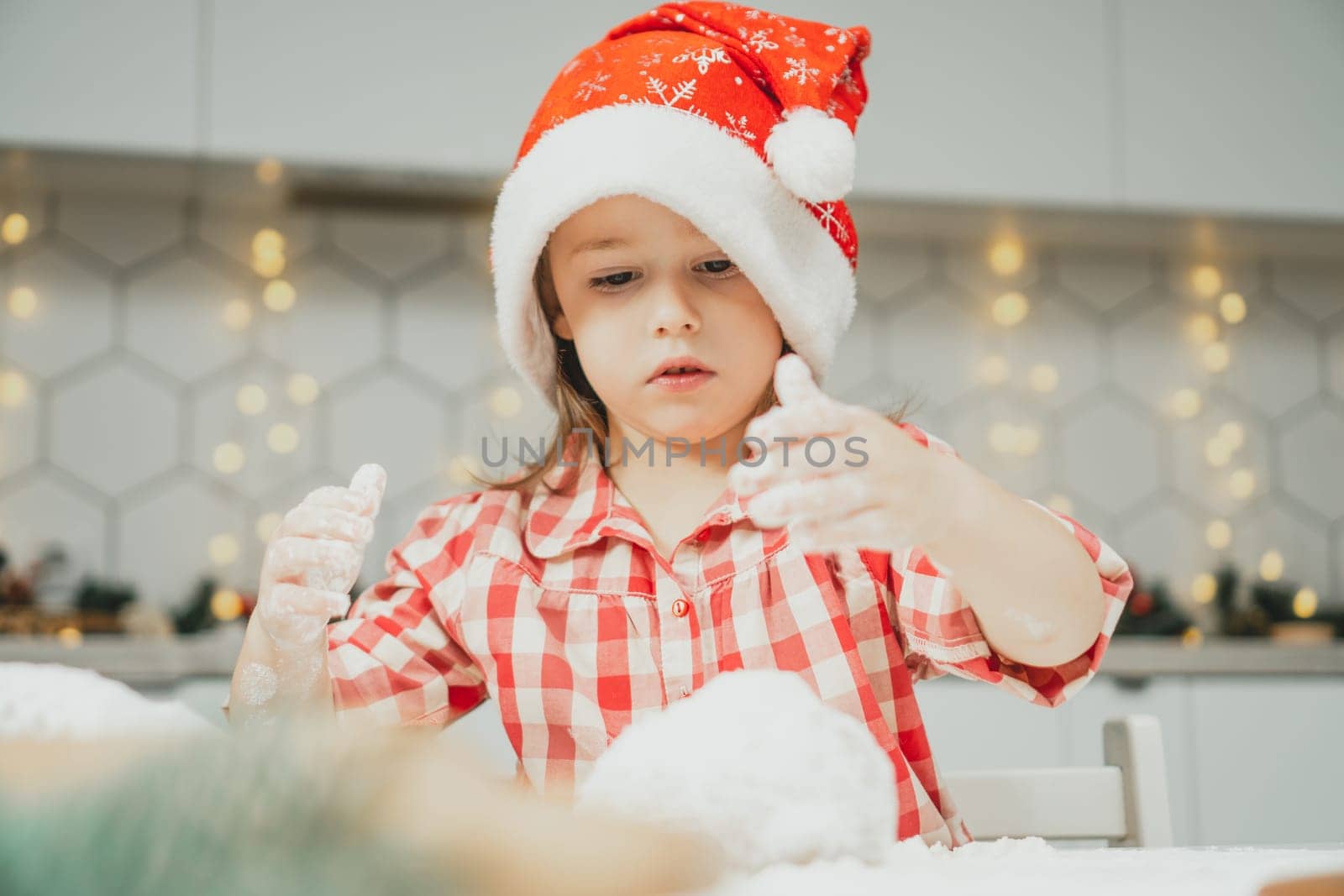 Little dark-haired girl 3 years old in red Christmas cap and checkered shirt prepares dough for gingerbread cookies in white Christmas decorated kitchen with garland lights. Merry Christmas, New Year