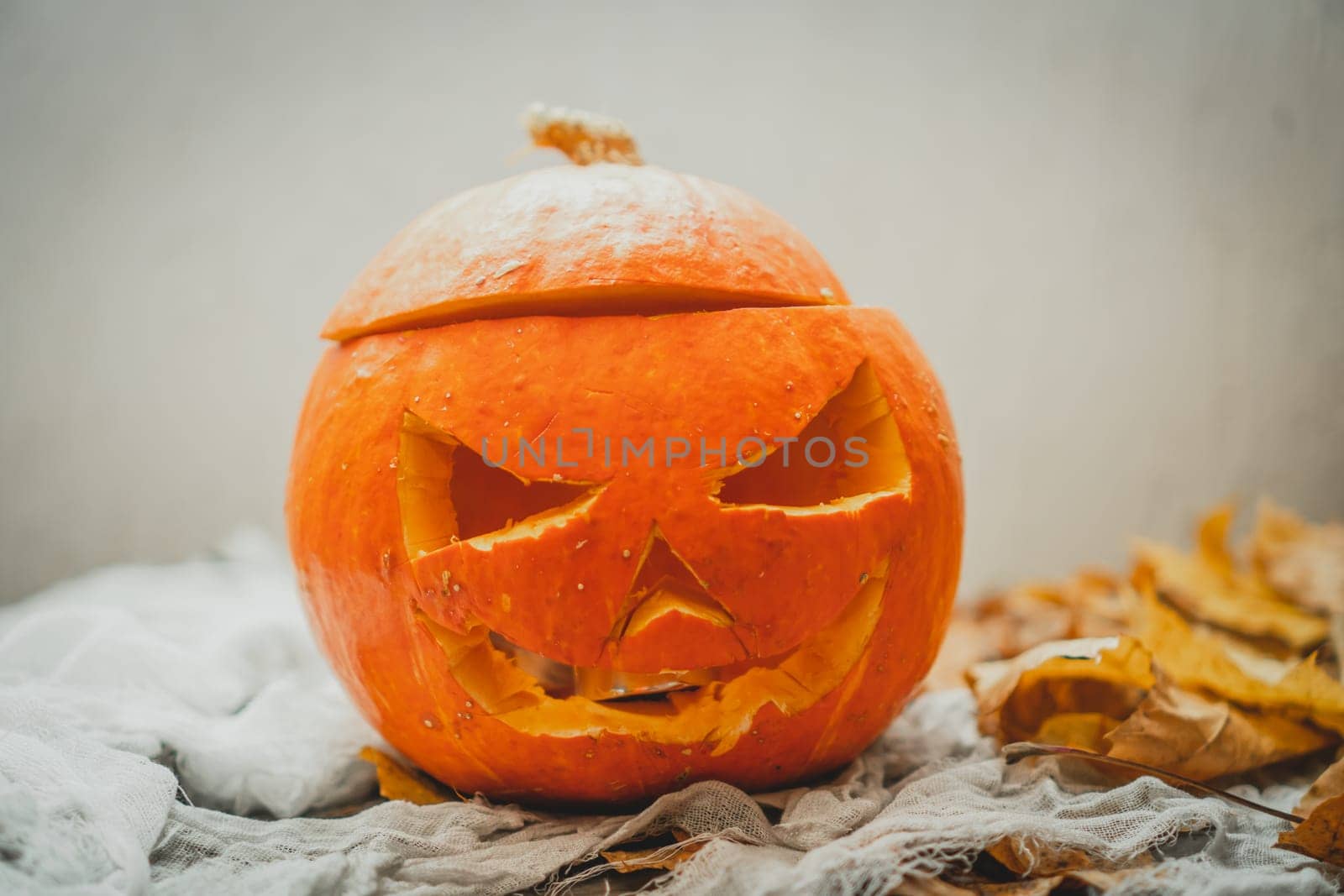Orange bright halloween pumpkin with carved nose, mouth and eyes on light gray background next to autumn yellow leaves. Scary face carved on vegetable. Halloween holiday decoration