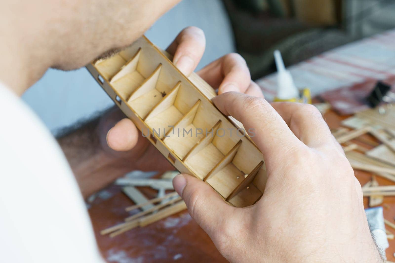 Hands of man gluing plywood details for ship model with glue, holding with fingers. Process of building toy ship, hobby, handicraft. Table with various materials, parts and devices for work