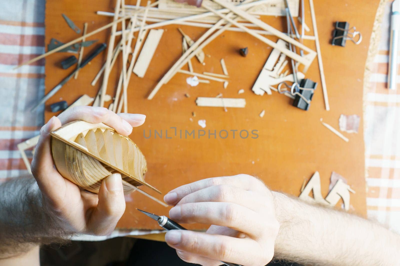 Hands of man removing excess glue from plywood details for ship model with utility knife. Process of building toy ship, hobby, handicraft. Table with various materials. Top view