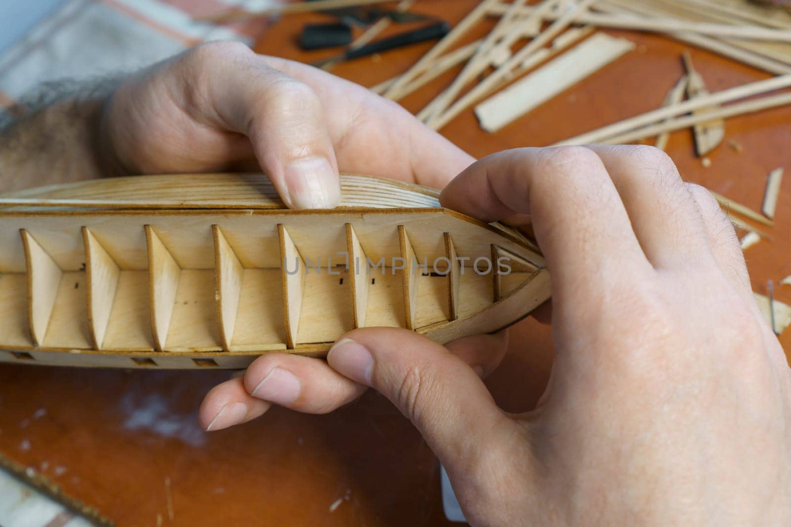 Hands of man gluing plywood details for ship model with glue, holding with fingers. Process of building toy ship, hobby, handicraft. Table with various materials, parts and devices for work