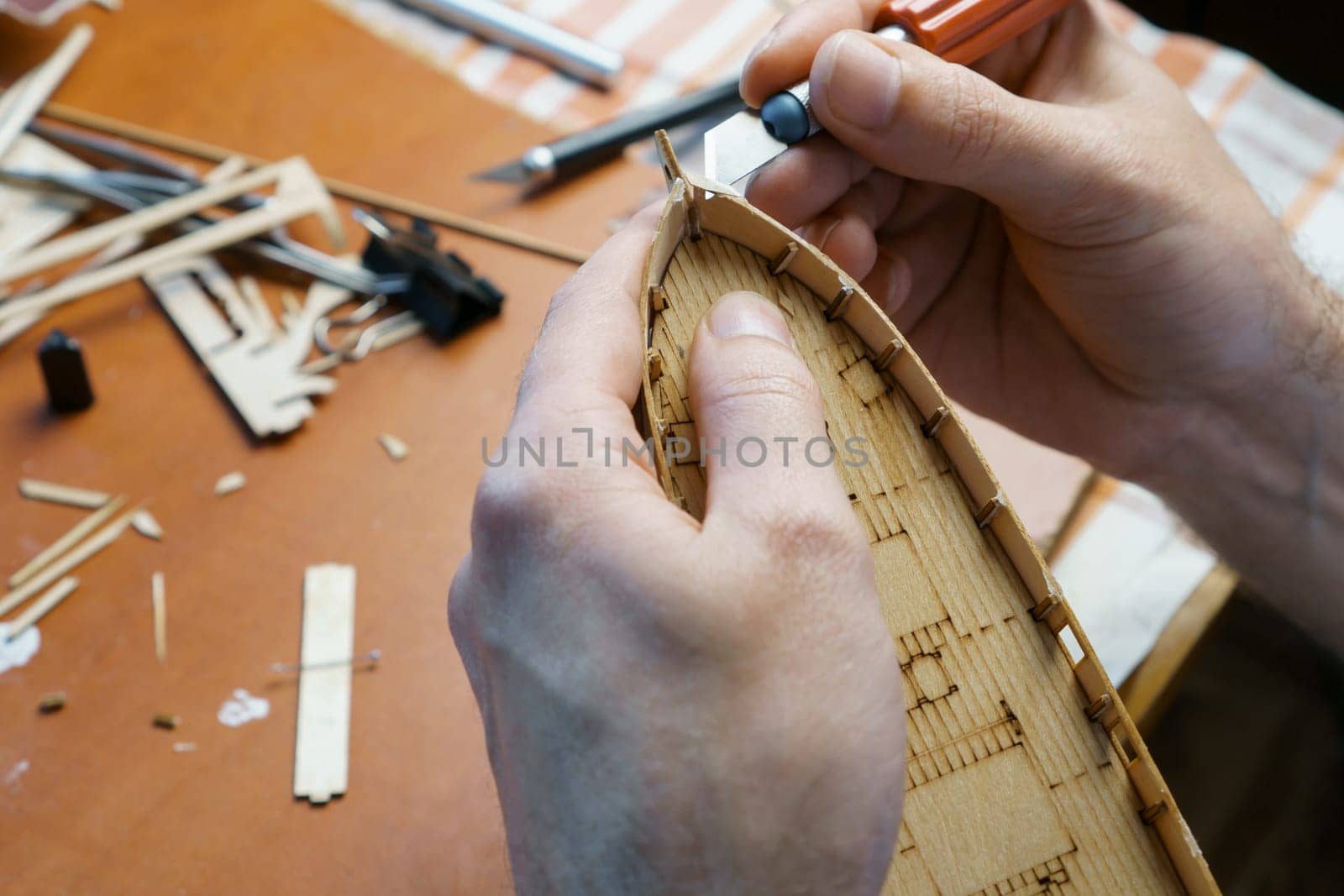 Hands of man cutting out details for ship model with clerical knife from plywood. Process of building toy ship, hobby and handicraft. Table with various materials, parts and devices for work