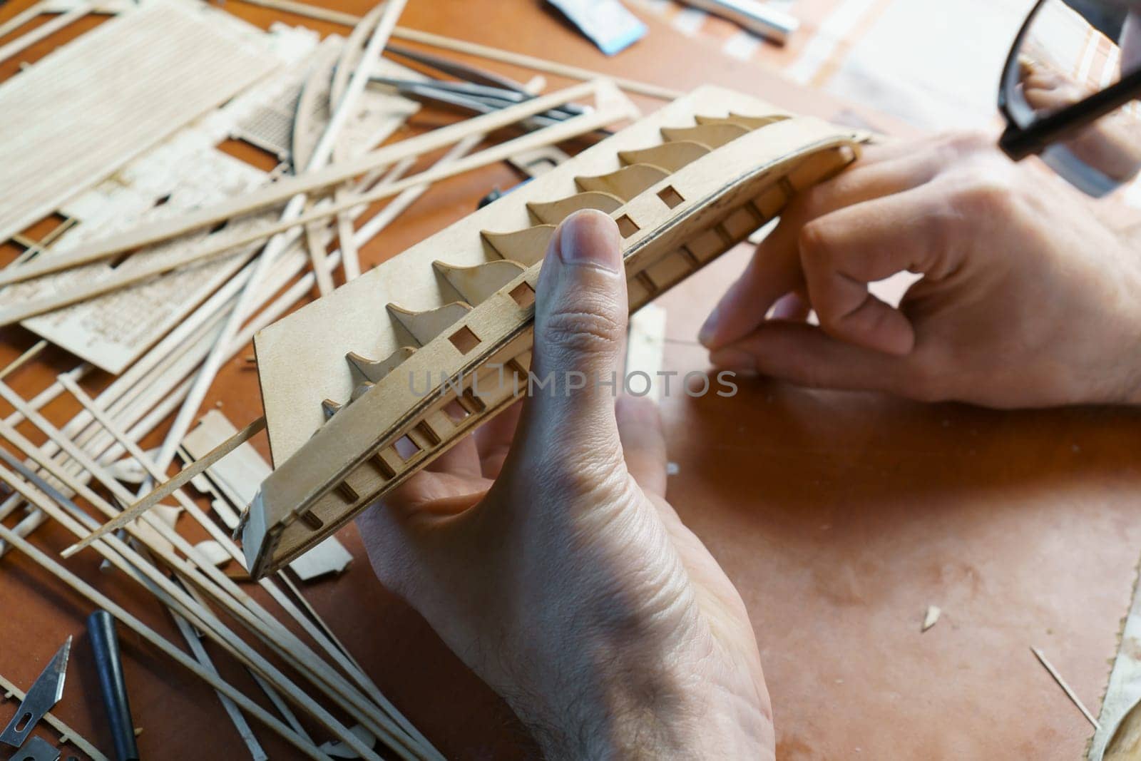 Hands of man gluing plywood details for ship model with glue, holding with fingers. Process of building toy ship, hobby, handicraft. Table with various materials, parts and devices for work