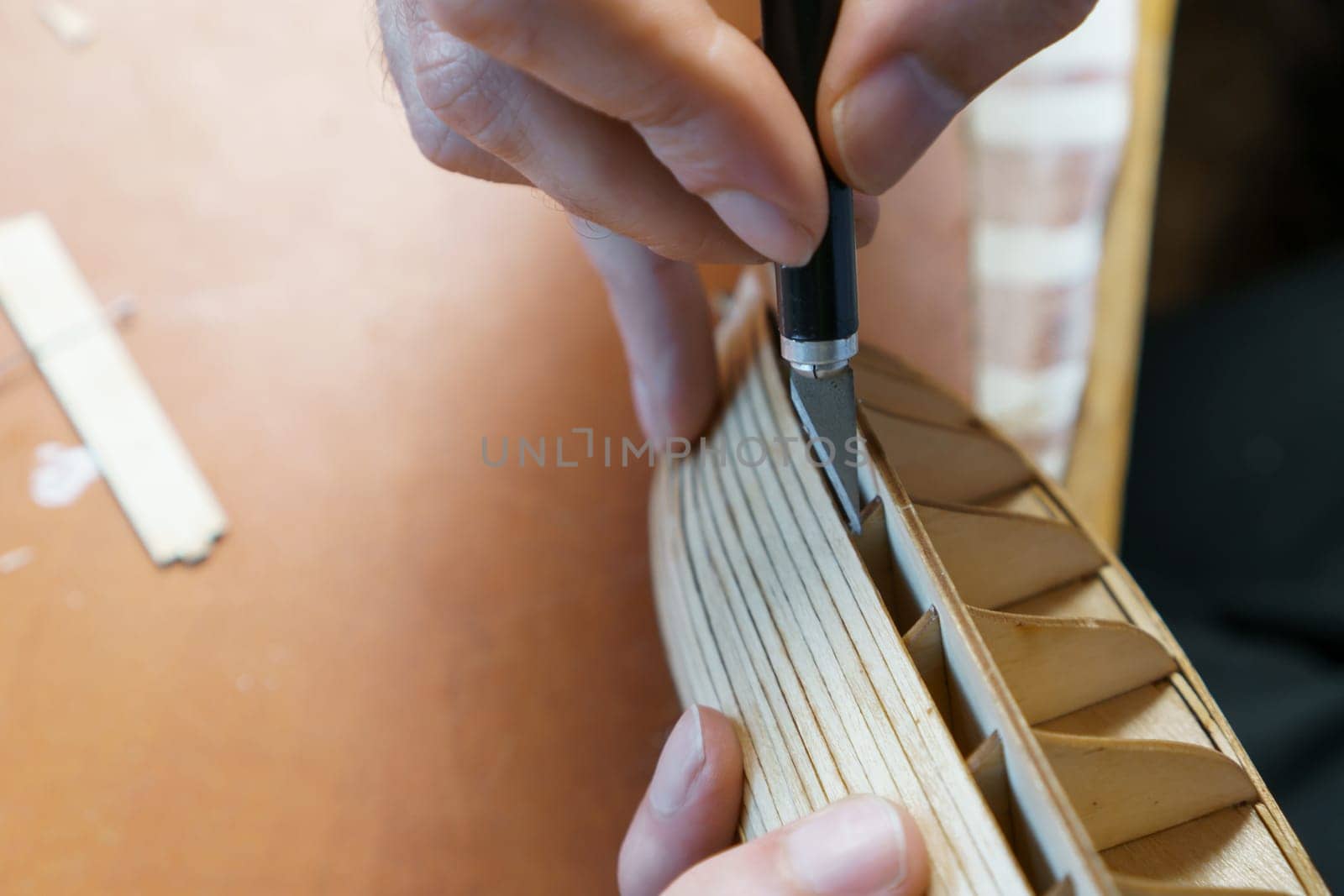 Hands of man removing excess glue from plywood details for ship model with utility knife. Process of building toy ship, hobby, handicraft. Table with various materials