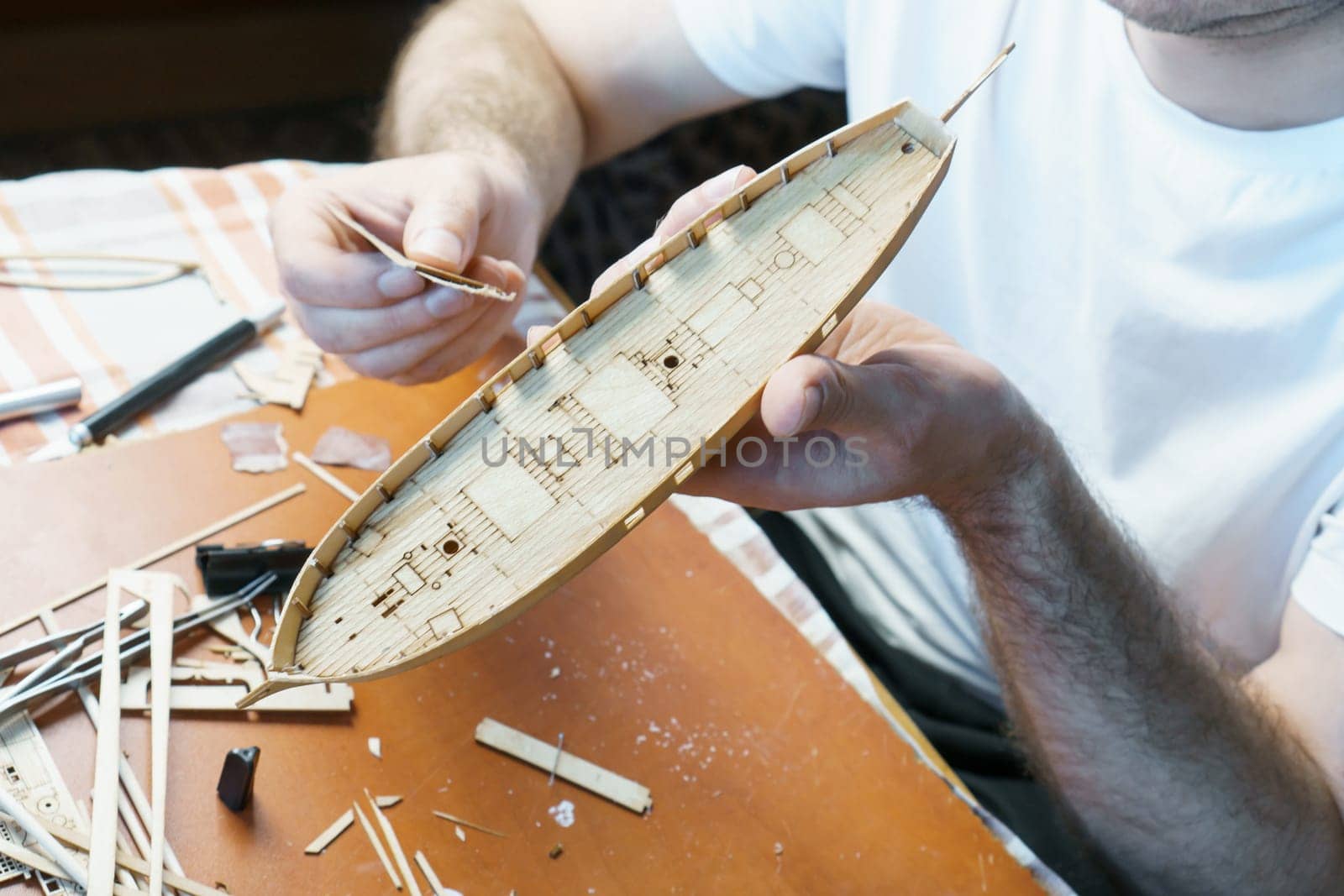Hands of man adjusts plywood details for ship model, grinding on sandpaper. Process of building toy ship, hobby and handicraft. Table with various materials, parts and devices for work