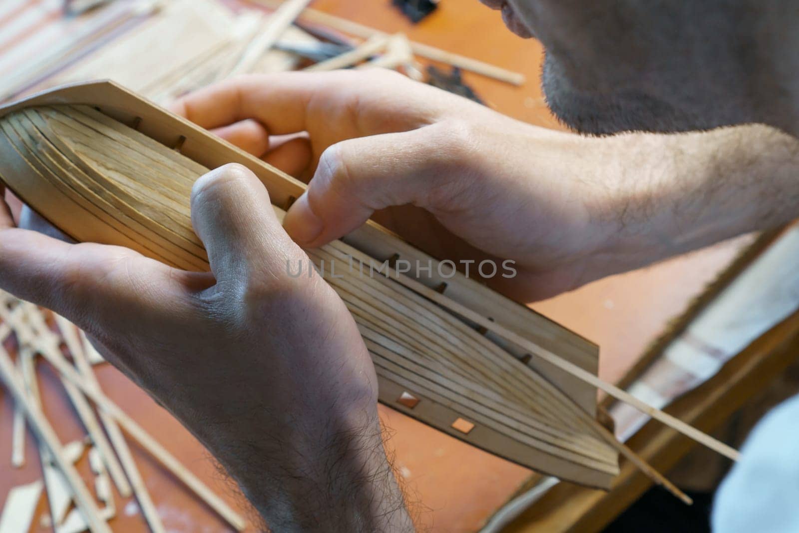 Hands of man gluing plywood details for ship model with glue, holding with fingers. Process of building toy ship, hobby, handicraft. Table with various materials, parts and devices for work