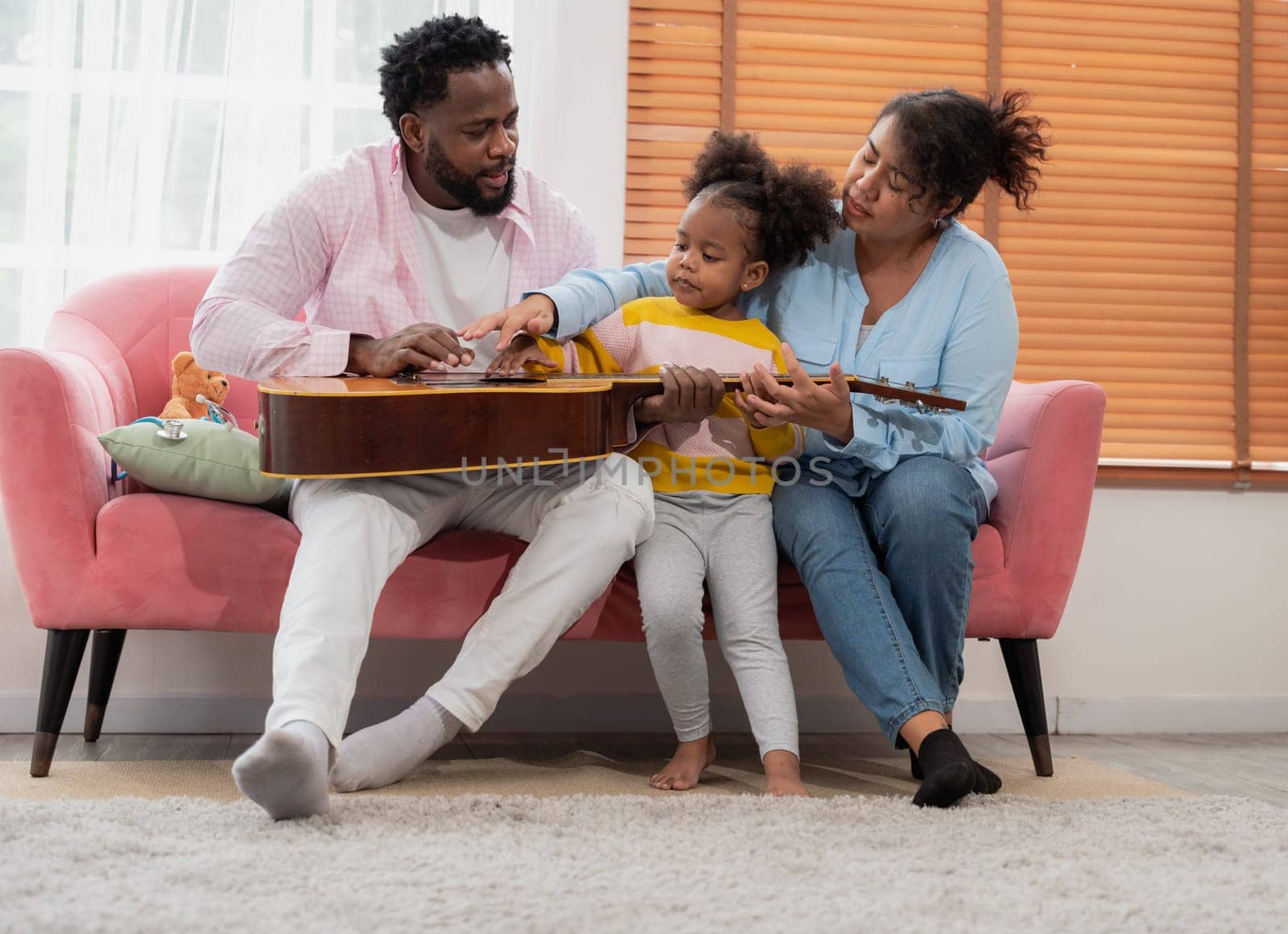 Happy african family with little curly girl sitting relax on pink couch and playing guitar together in living room at home
