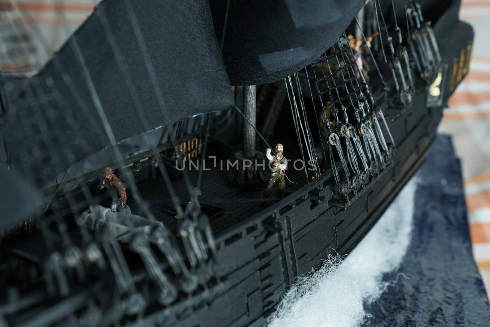 Pirate ship model Black pearl made of plywood, with miniature figures of pirate sailors, close-up details. Ship sails on wave. Sailors on deck pulling ropes, partially blurred. Patched sails belly out