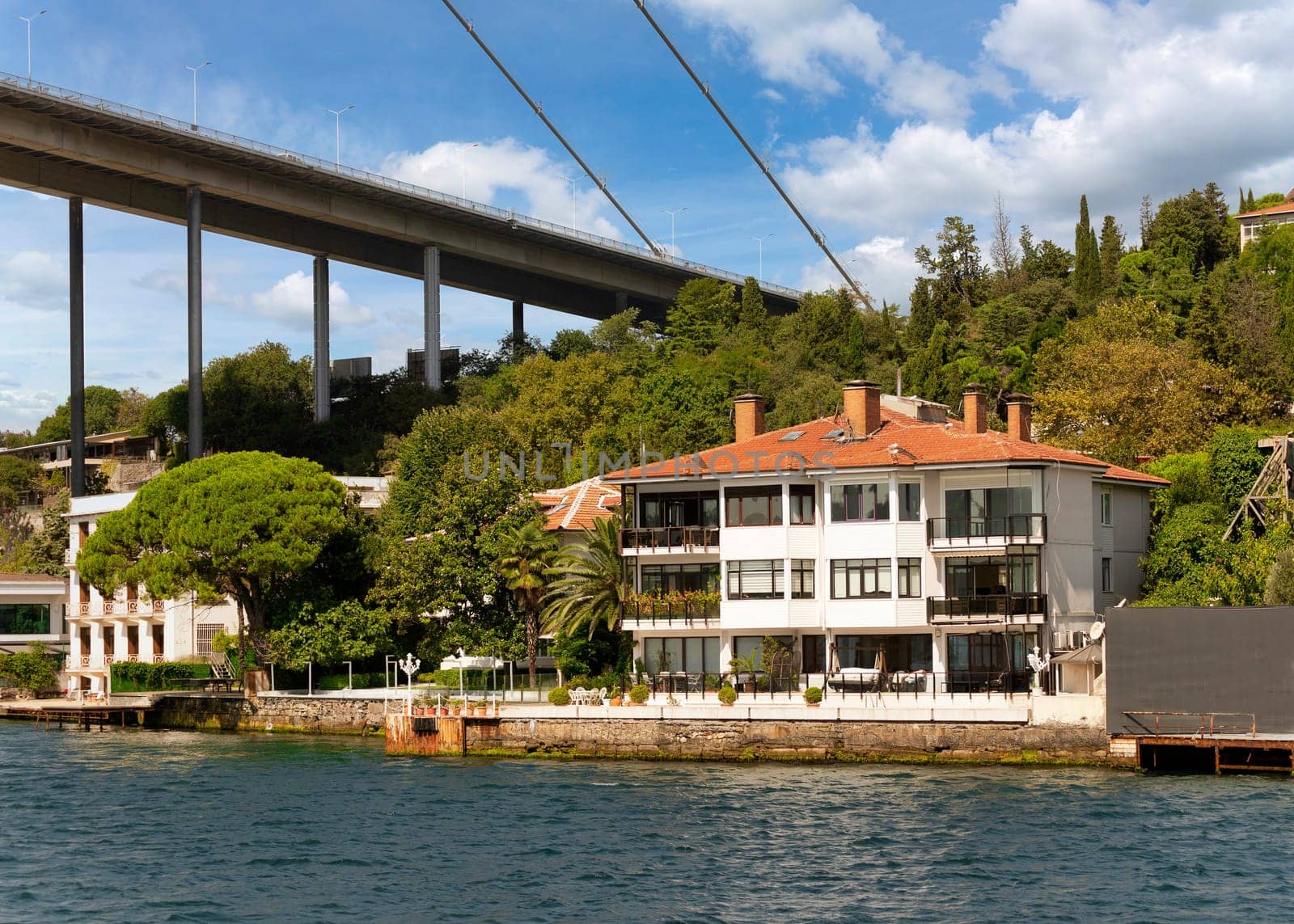 View from the sea of the European side of Bosphorus strait, Istanbul, Turkey, with traditional waterfront house, under Bosphorus bridge, and green hill with dense trees, in a summer day