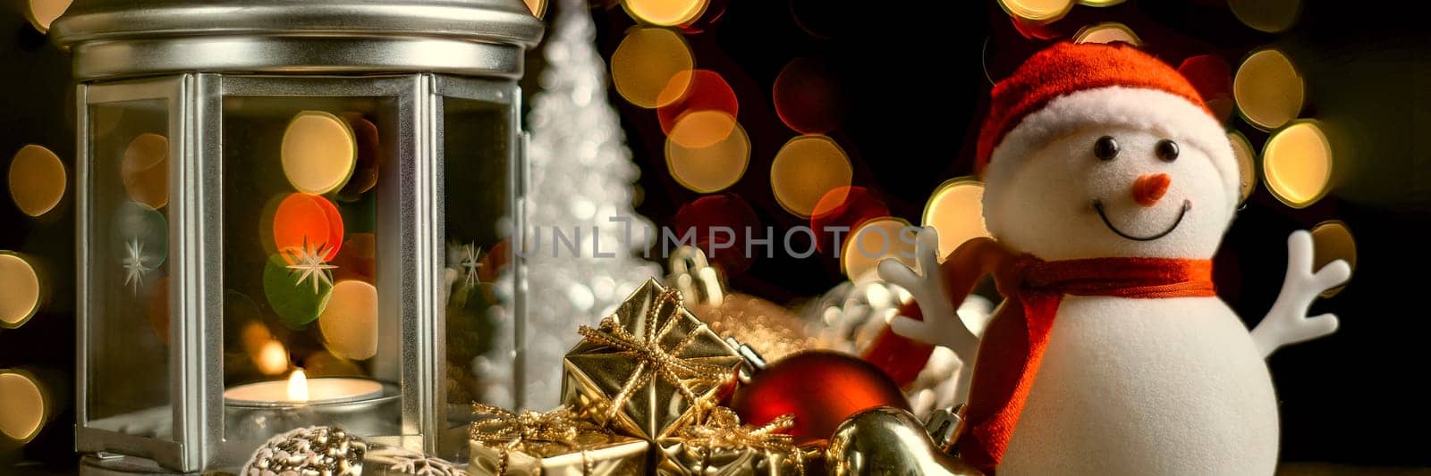 Christmas composition: A snowman, a lantern with candles, bright shiny boxes with gifts, blurred bokeh lights in the background.