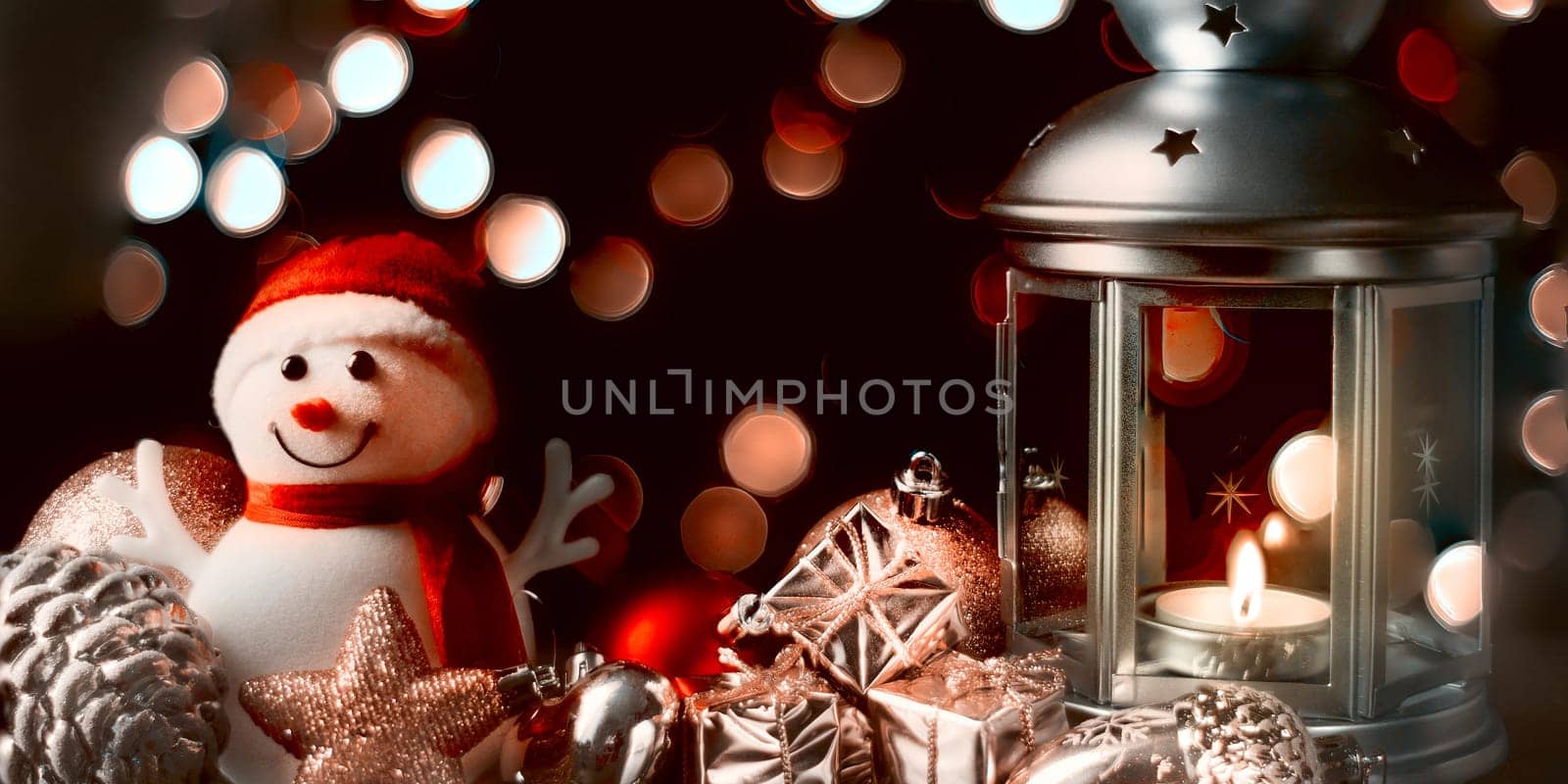 Christmas composition: A snowman, a lantern with candles, bright shiny boxes with gifts, blurred bokeh lights in the background.