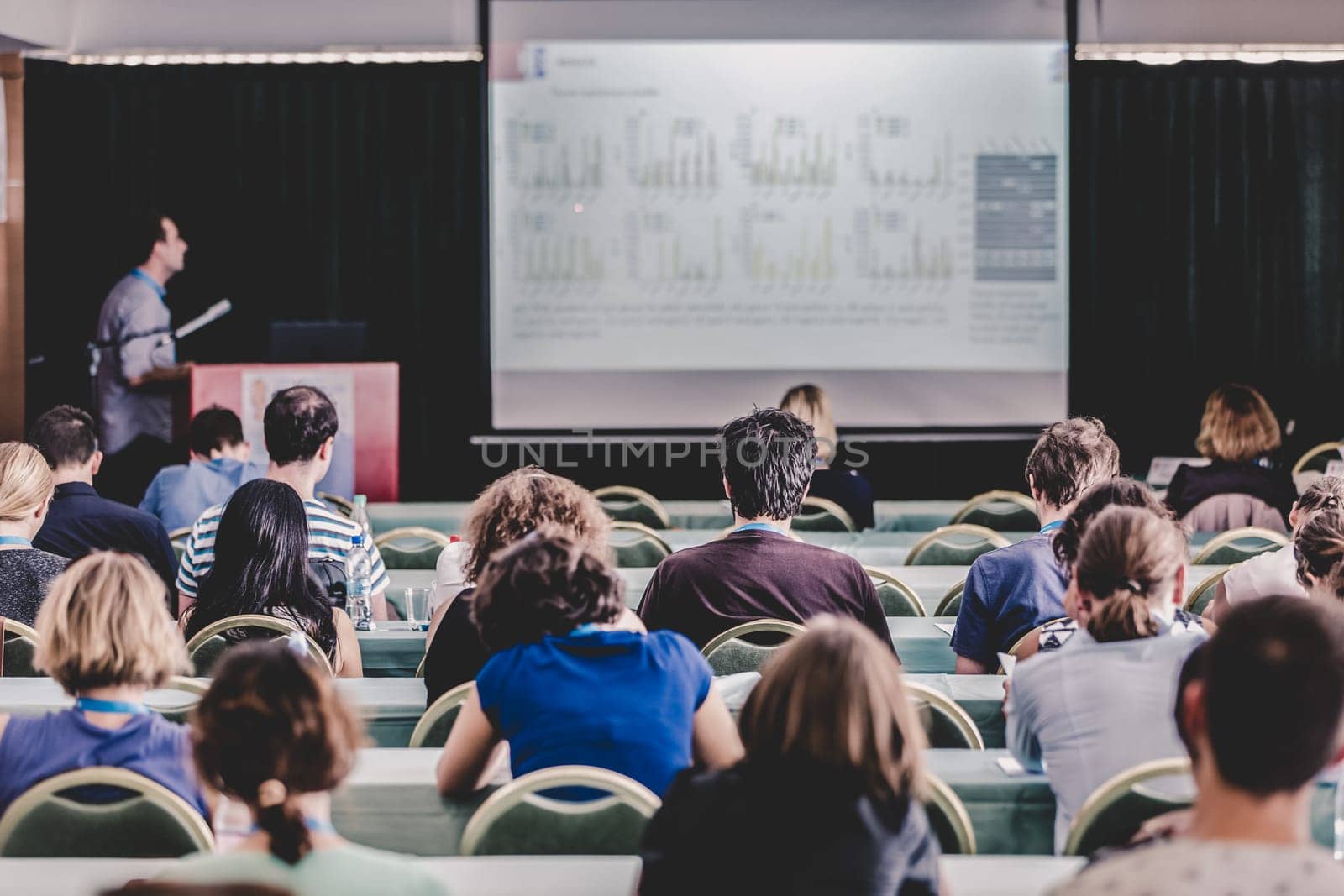 Speaker Giving a Talk at Business Meeting. Audience in the conference hall. Business and Entrepreneurship. Focus on unrecognizable people from rear.